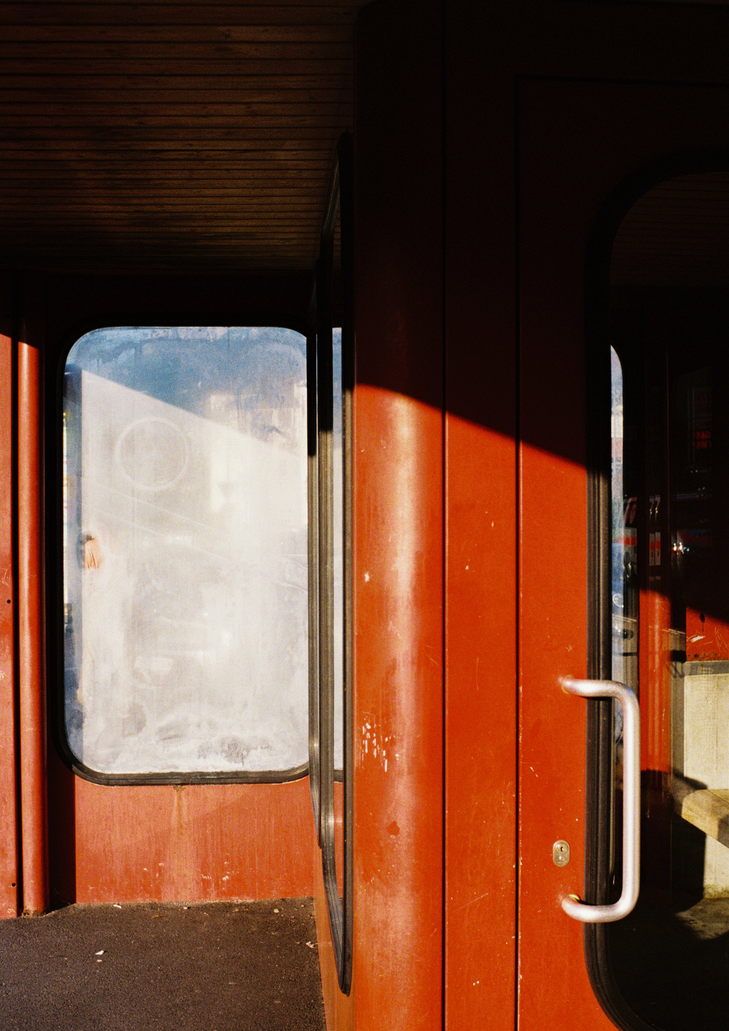Close-up of a building entrance with red walls and glass doors, one door slightly open, showing stairs inside and a reflection of the street in the glass.