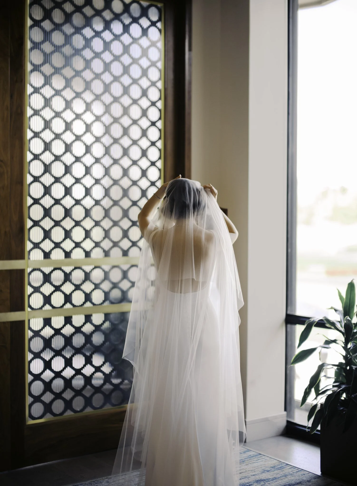 A woman in a wedding dress and veil standing by a window, adjusting her hair or veil indoors.