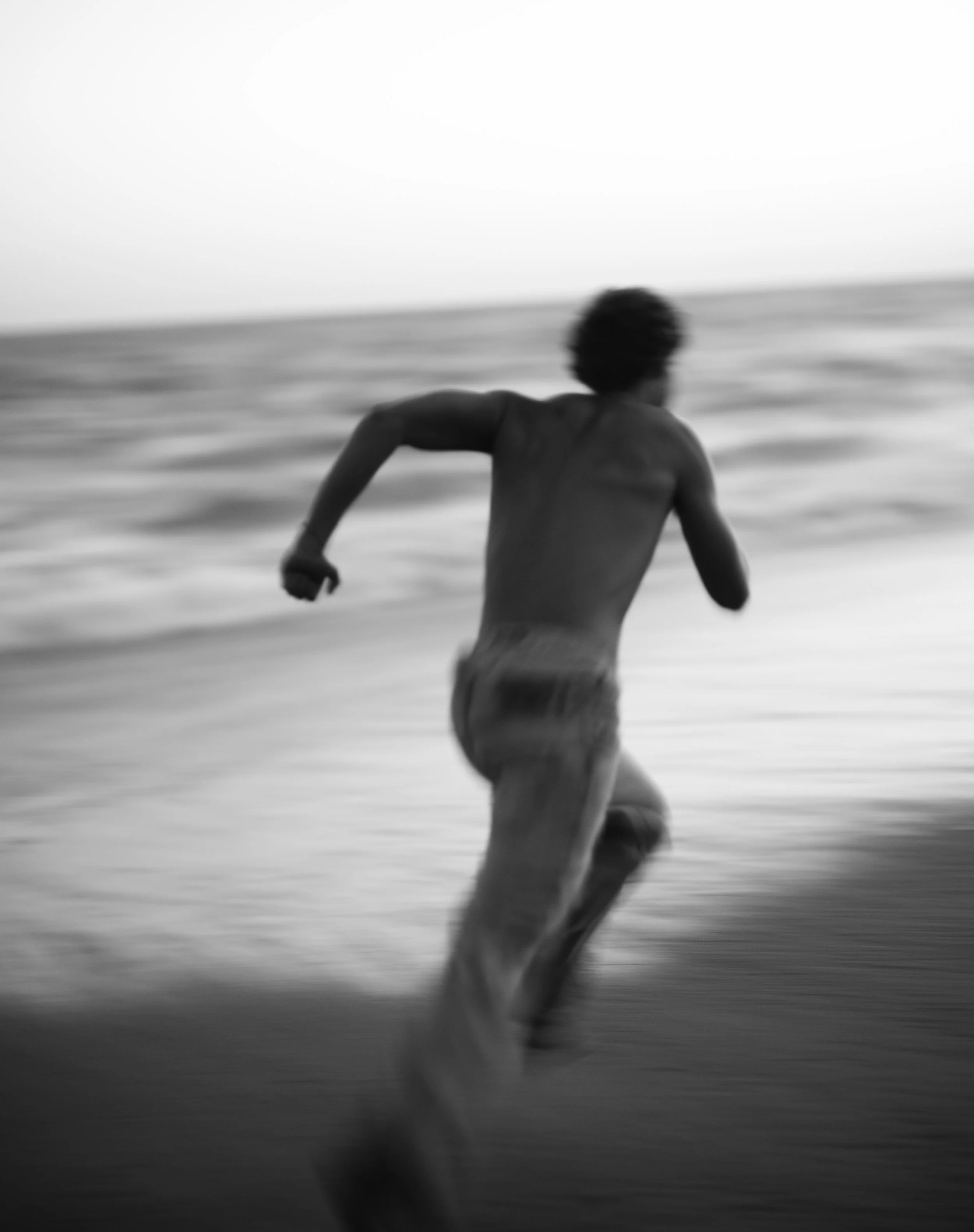 Black and white photograph of a man running into the ocean, seen from behind, with blurred motion and waves in the background.