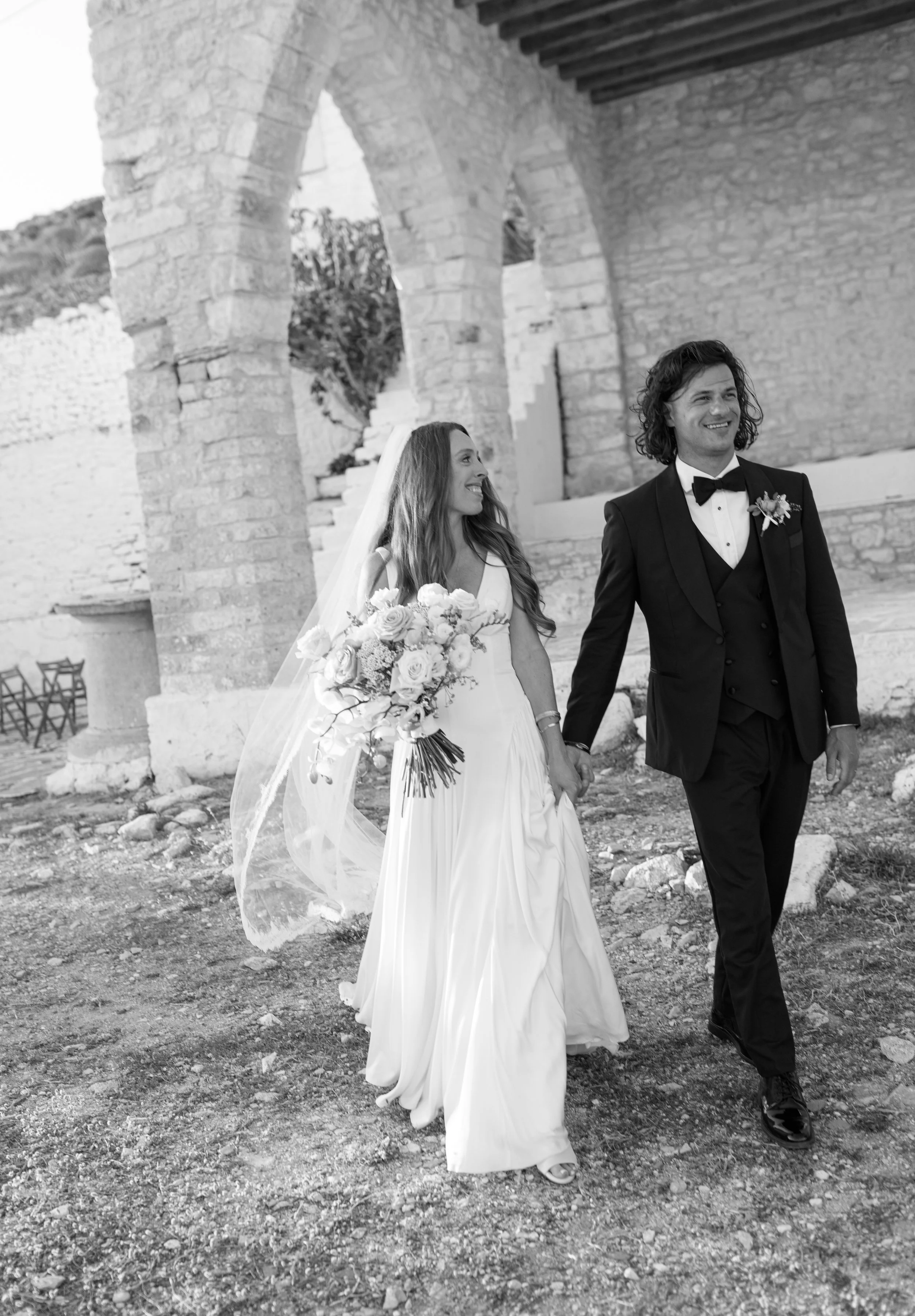 A bride and groom walking hand-in-hand outdoors near Roman-style stone arches. The bride is holding a bouquet of flowers, wearing a white dress and veil, smiling at the groom who is dressed in a tuxedo with a bow tie, also smiling.