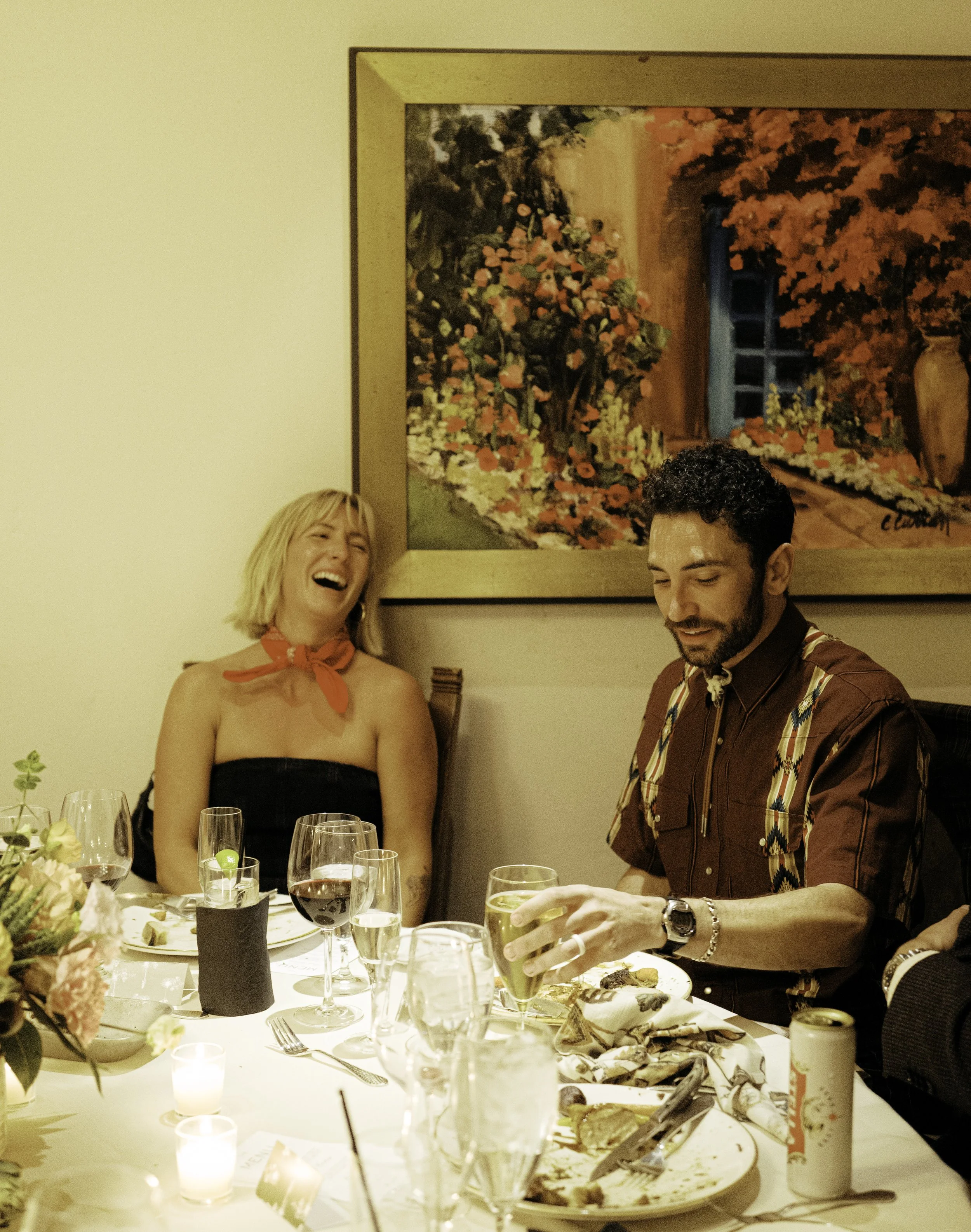 A joyful blonde woman and a man with dark hair and beard, sitting at a dining table with glasses of wine and other beverages, celebrating in a warmly lit room with a floral arrangement and a large framed painting of a rustic scene on the wall behind 