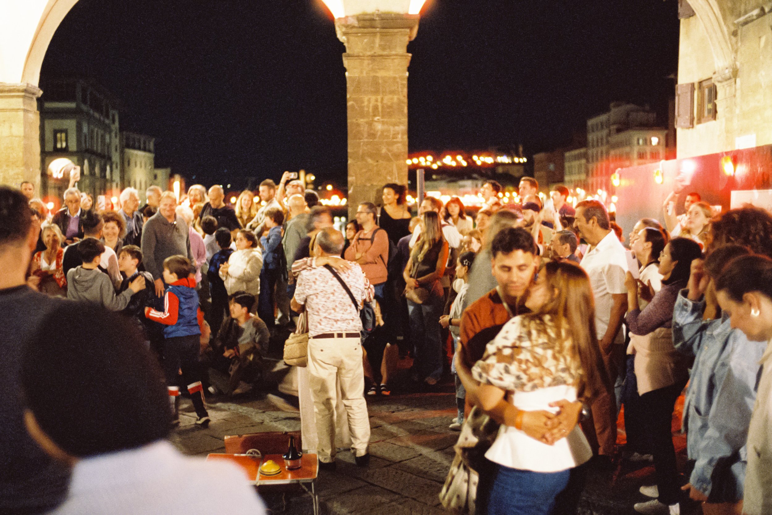 A crowded nighttime scene under a stone arch, with people gathered on a city street, some taking photos, others talking or walking, illuminated by streetlights and building lights.