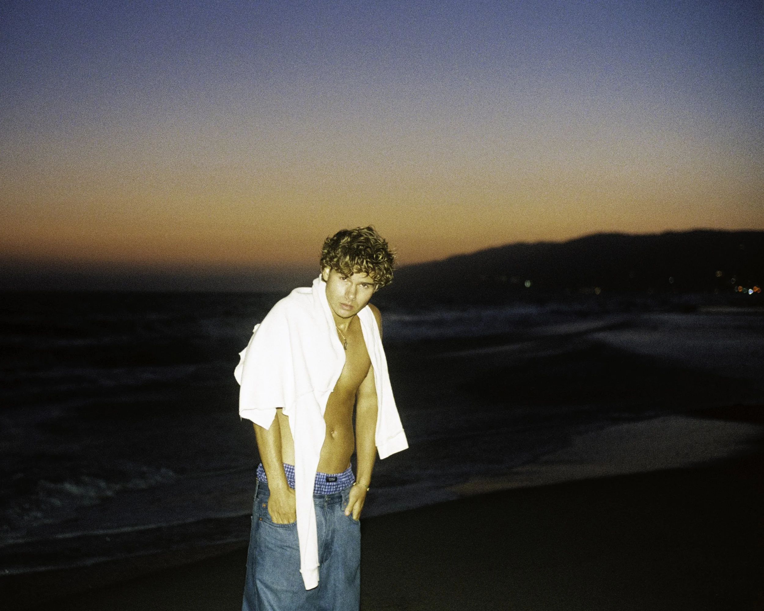 Young man with curly hair at the beach during sunset, wearing jeans and an open shirt with a towel draped over his shoulders.