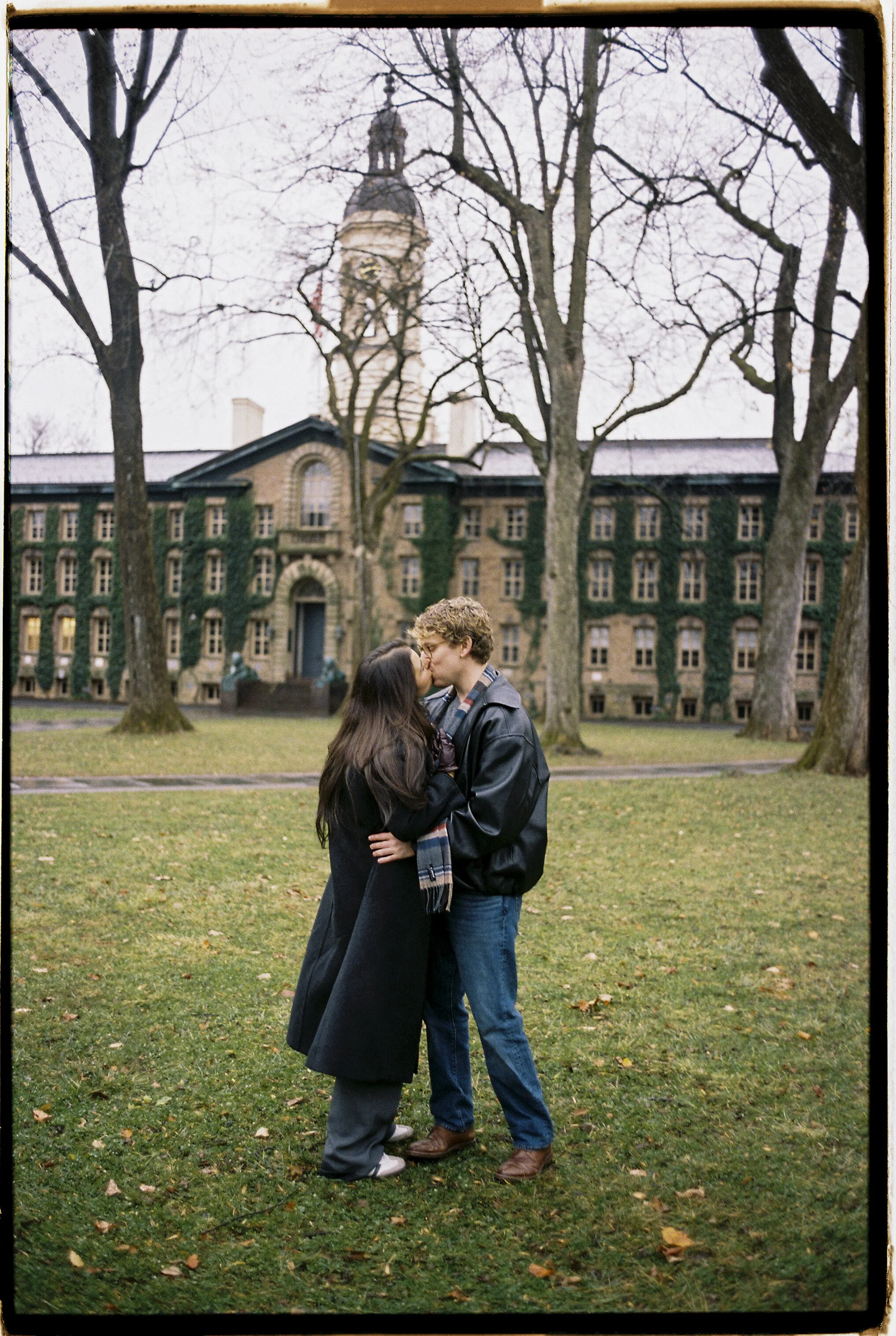 A couple kisses in a park with a large brick building and a clock tower in the background, surrounded by leafless trees and green grass.