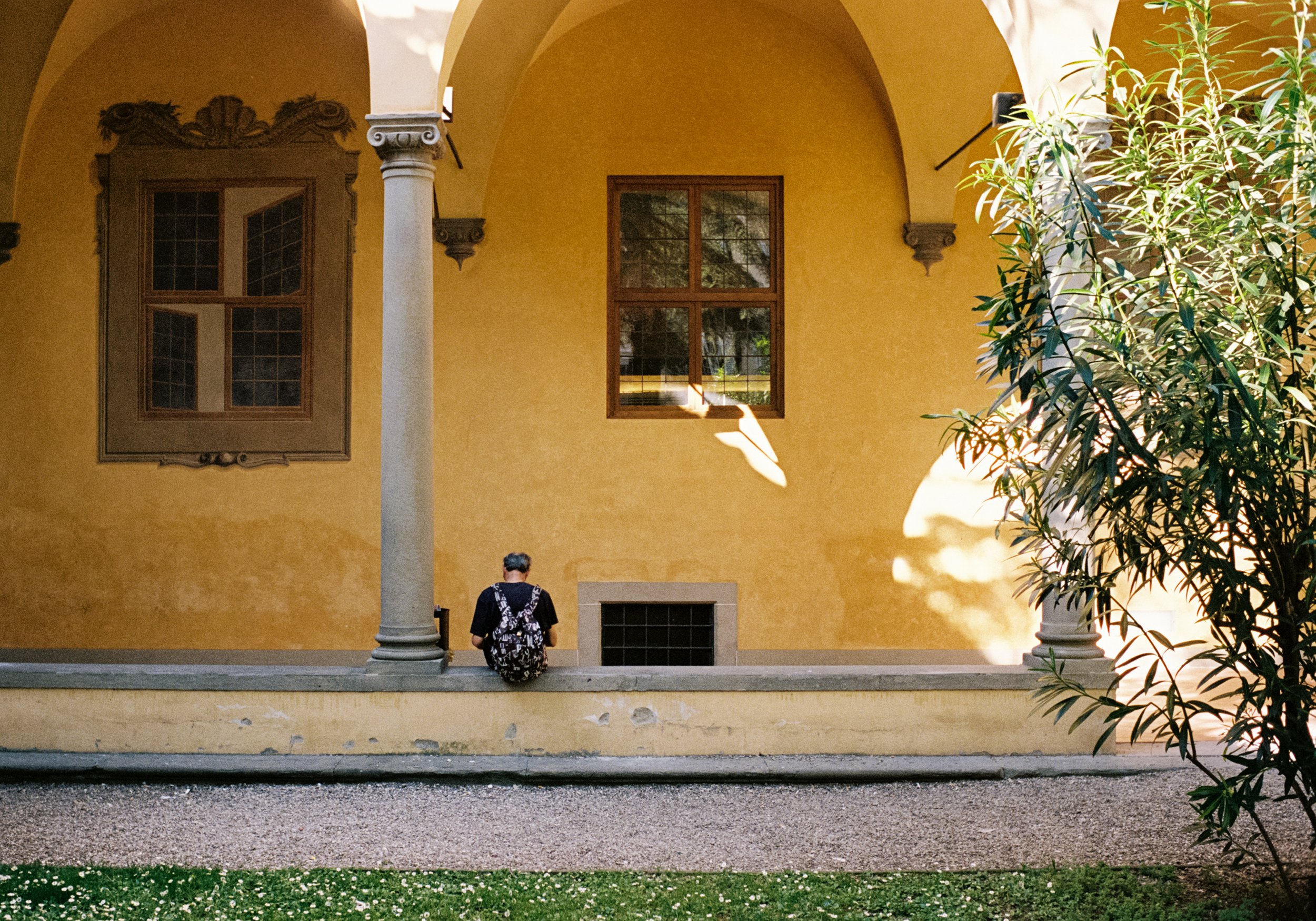 A person sitting on a low wall in front of a yellow building with decorative window frames and a large window. The person has their back to the camera and is wearing a black shirt and a floral backpack. Sunlight casts shadows on the yellow wall, and 