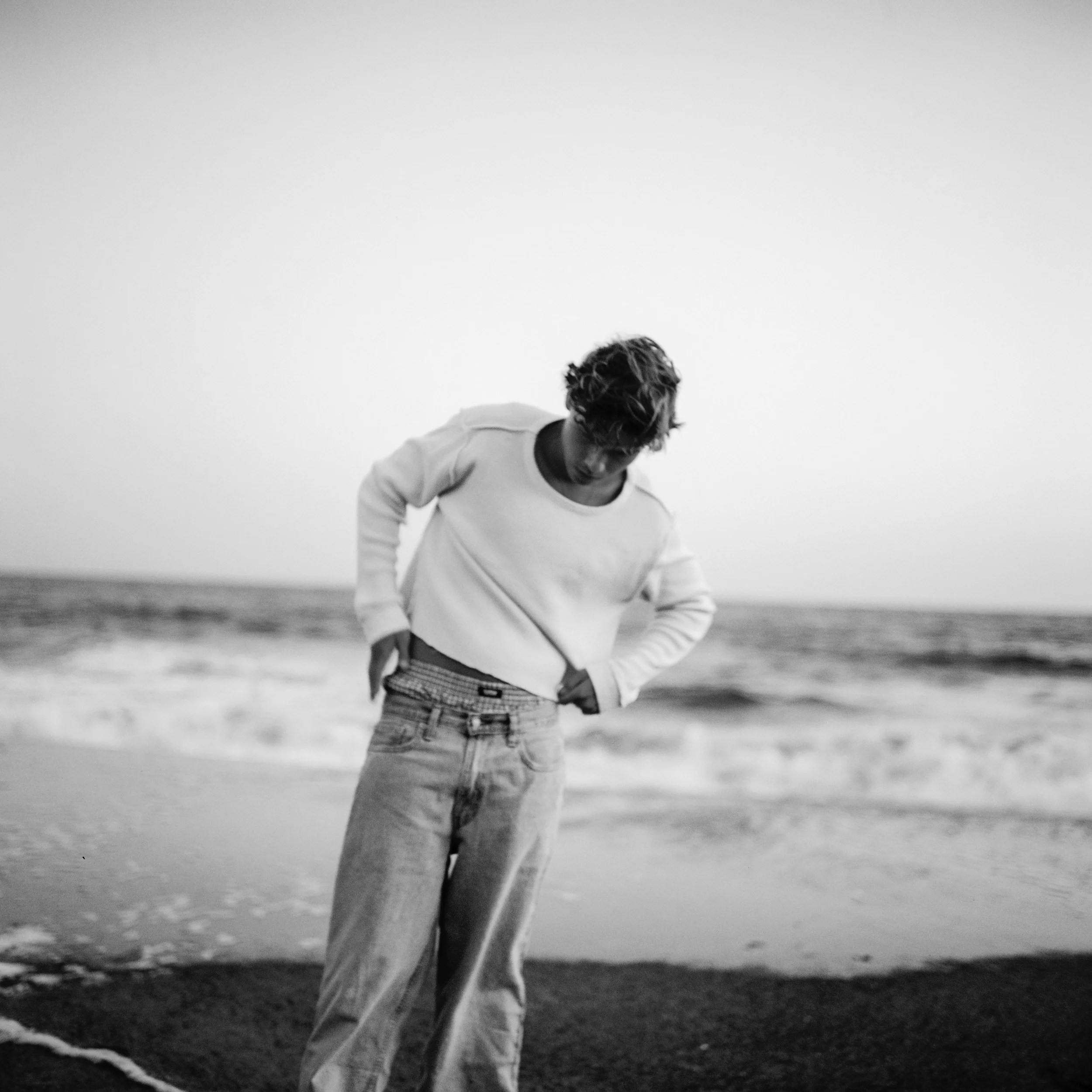 A person standing on a beach, looking down, with the ocean waves in the background, in black and white.