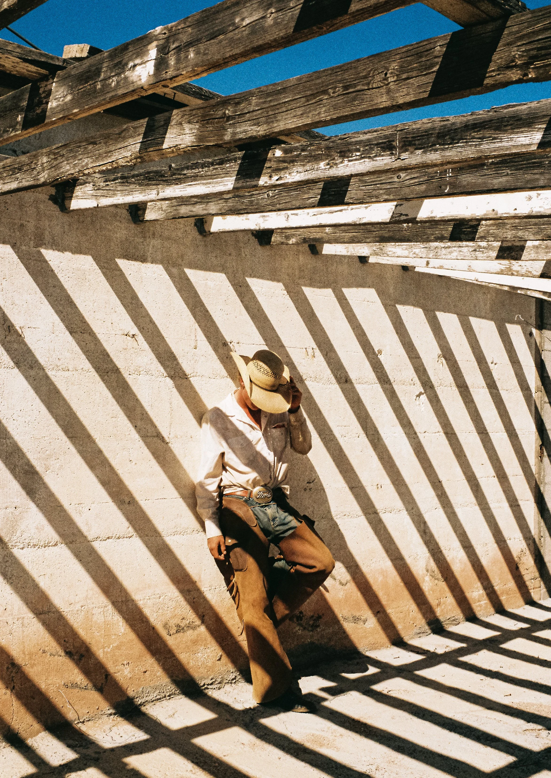 A person wearing a cowboy hat, white shirt, and brown pants is leaning against a concrete wall, talking on a phone, with striped shadows cast by a nearby wooden fence under a clear blue sky.