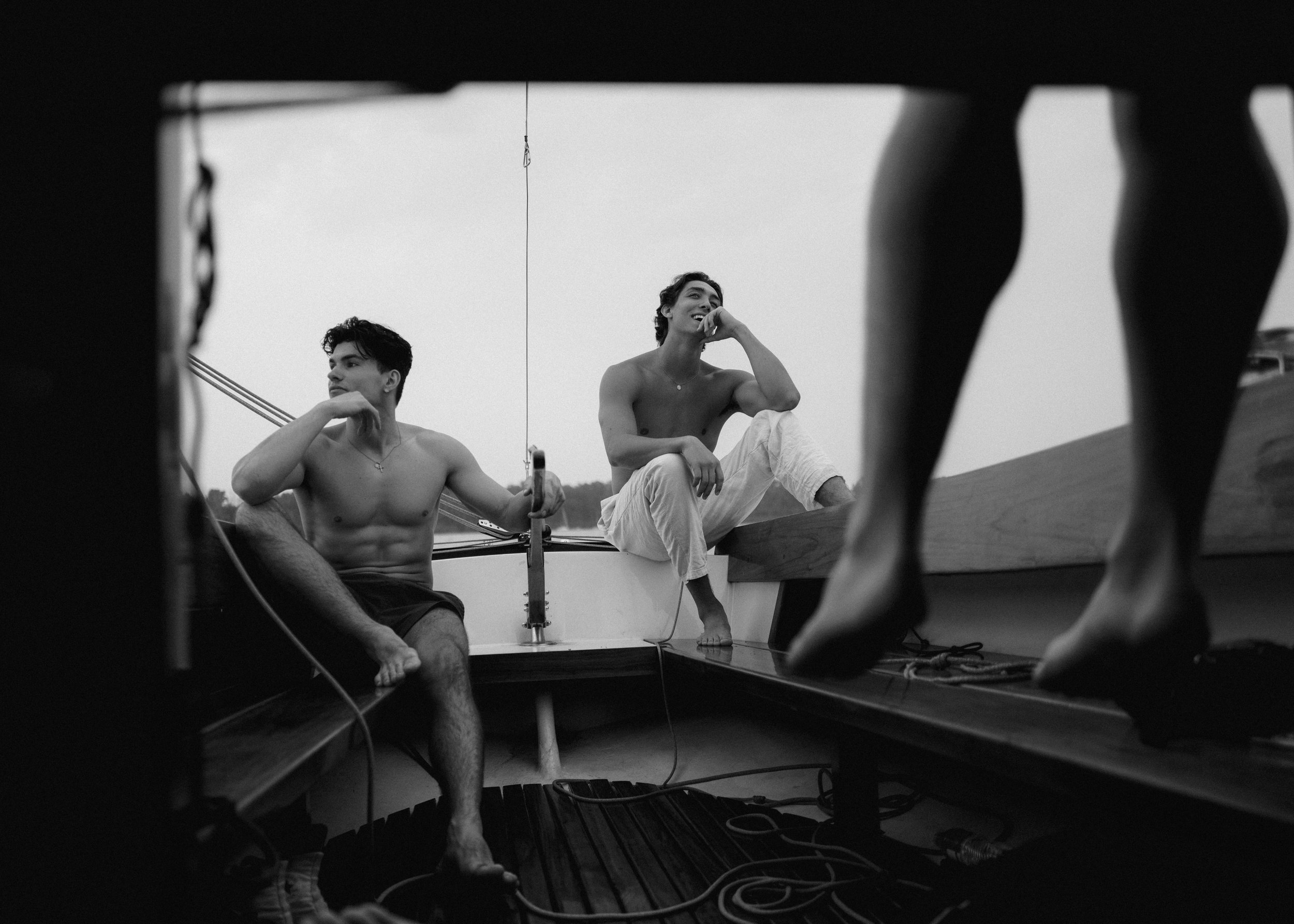 Two shirtless young men sitting on a boat, one looking into the distance, the other smiling and talking, seen through the legs of a person standing on the boat.