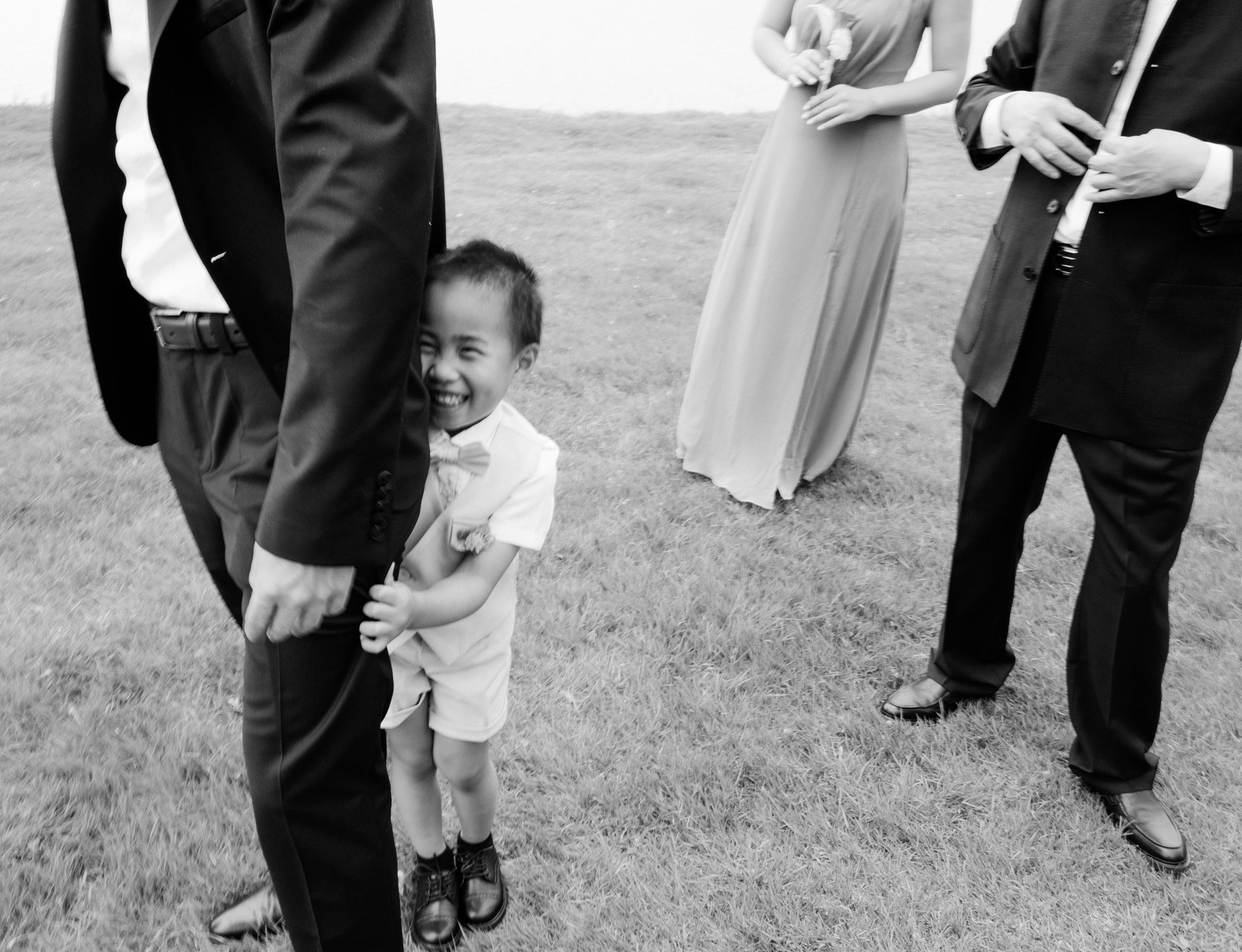 A group of adults and a child standing on grass, seen from the knees down, with some adults dressed in suits and a child in a light-colored outfit smiling and holding hands with someone.