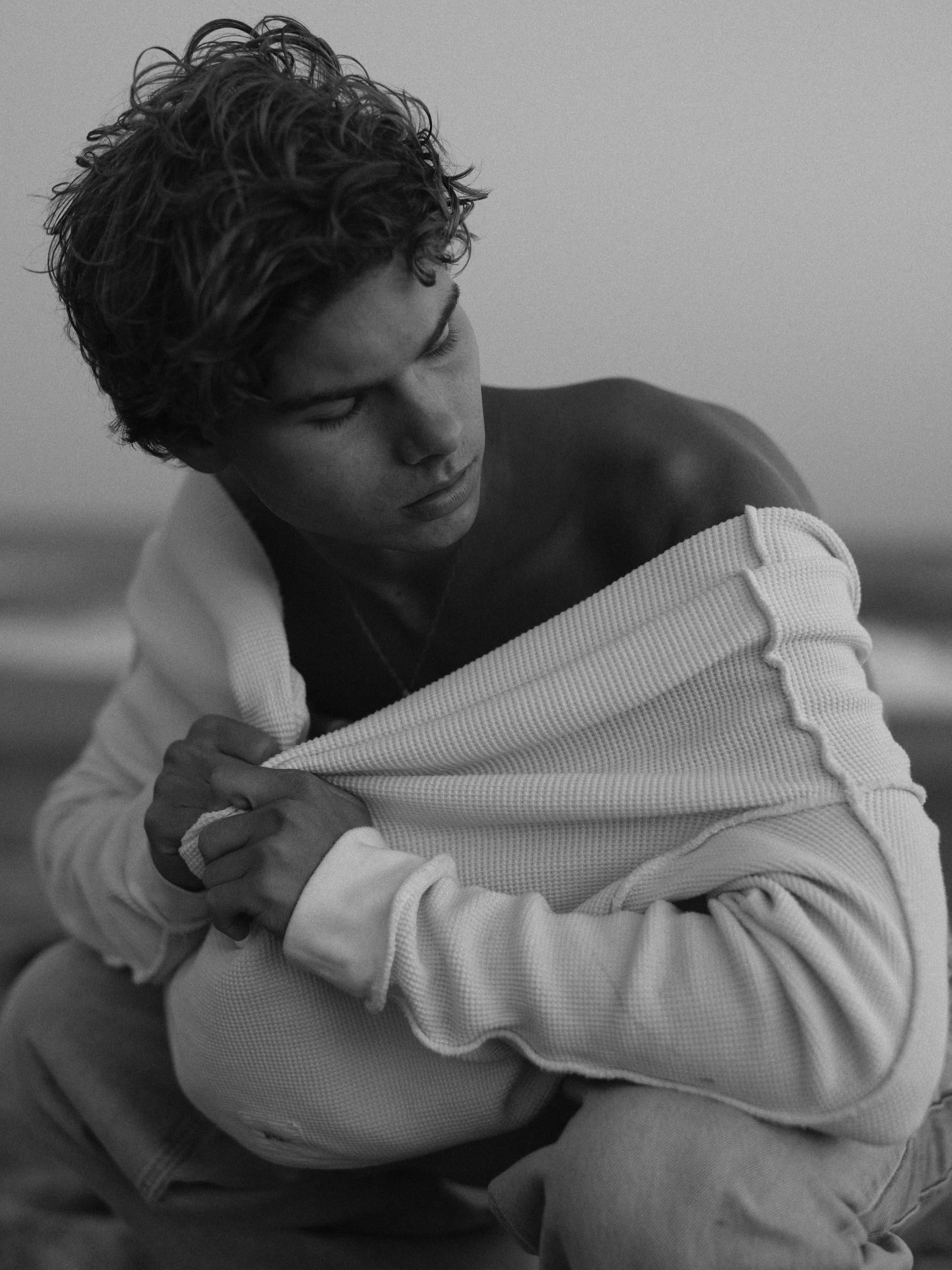 Black and white photo of a young man with curly hair, looking down while pulling off a white sweatshirt, exposing his shoulder and chest.