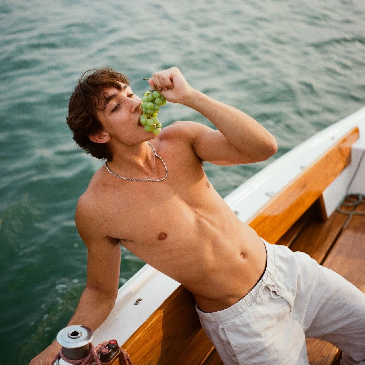 Young shirtless man on boat holding a bunch of green grapes near his mouth with water in the background.