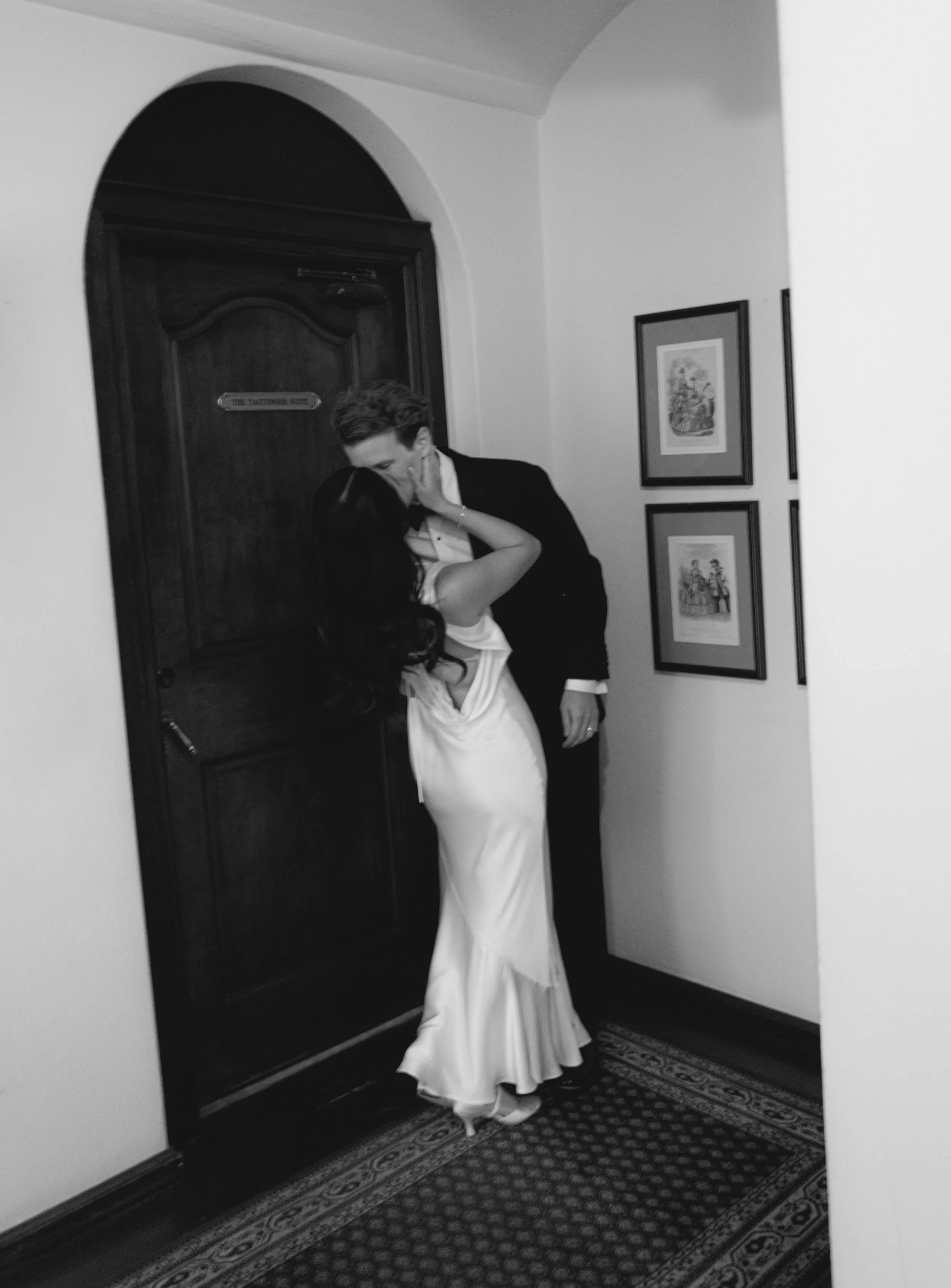 A couple kissing in front of a wooden door for the starting suite in a hallway. The woman is wearing a white dress, and the man is in a black suit.