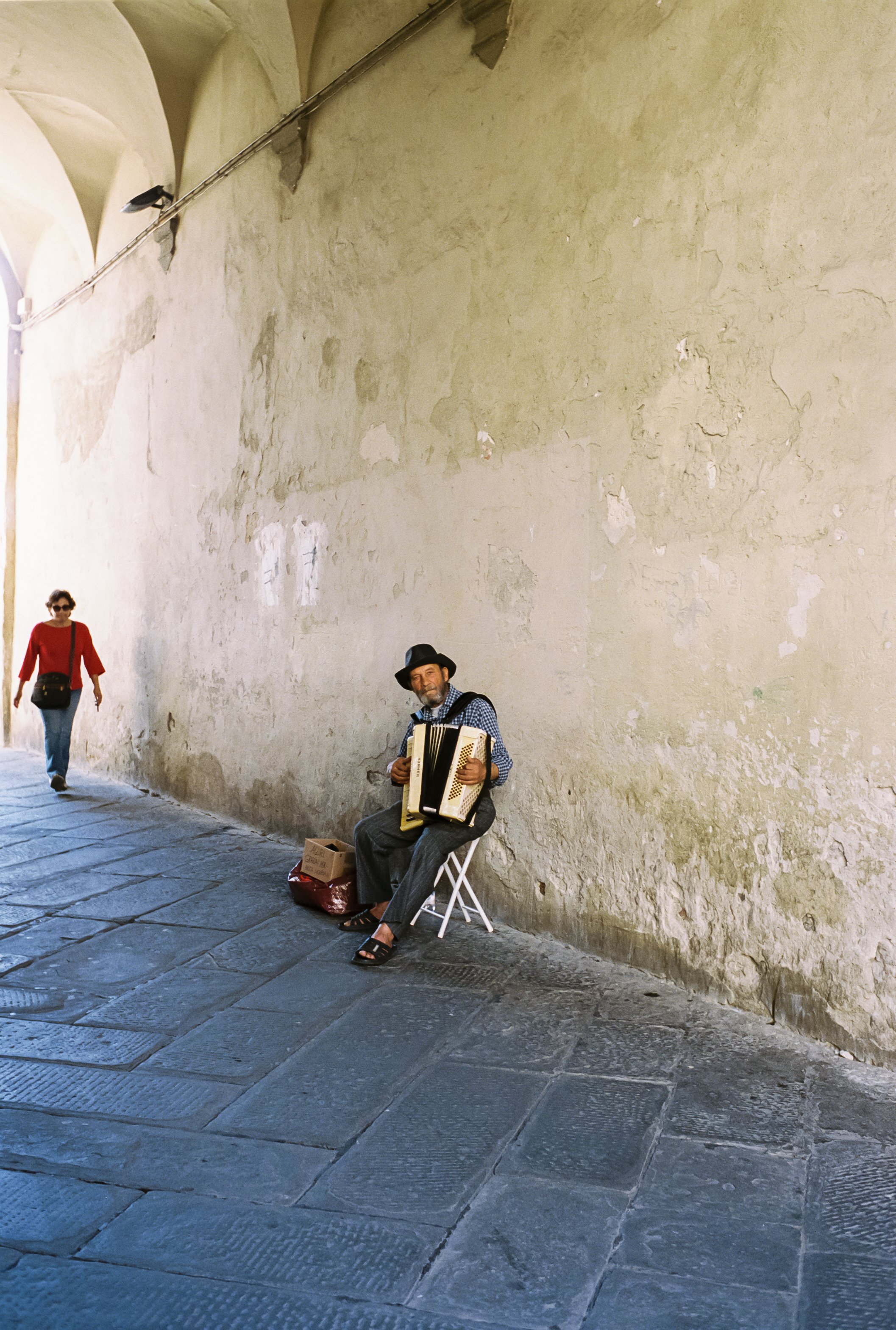 A street musician sitting on a small folding chair, playing an accordion. He is wearing a hat, a checkered shirt, and pants. There is a person walking away in the background, dressed in a red shirt and carrying a black bag.