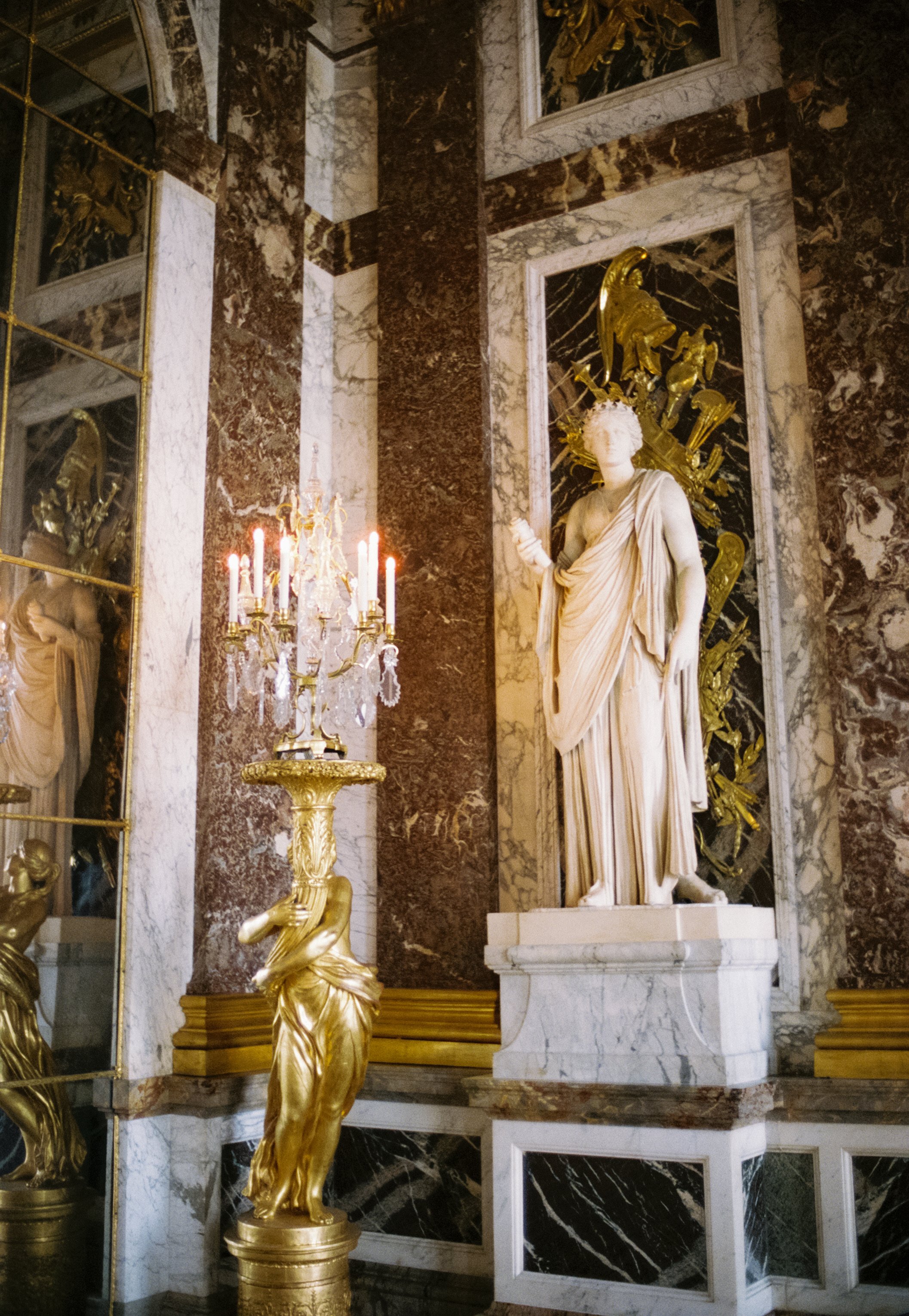 A marble statue of a draped woman holding an object in her hand, standing on a pedestal against a marble-paneled wall with gold accents. A gold and crystal chandelier with lit candles is in front of the statue, and a mirror to the left reflects part 
