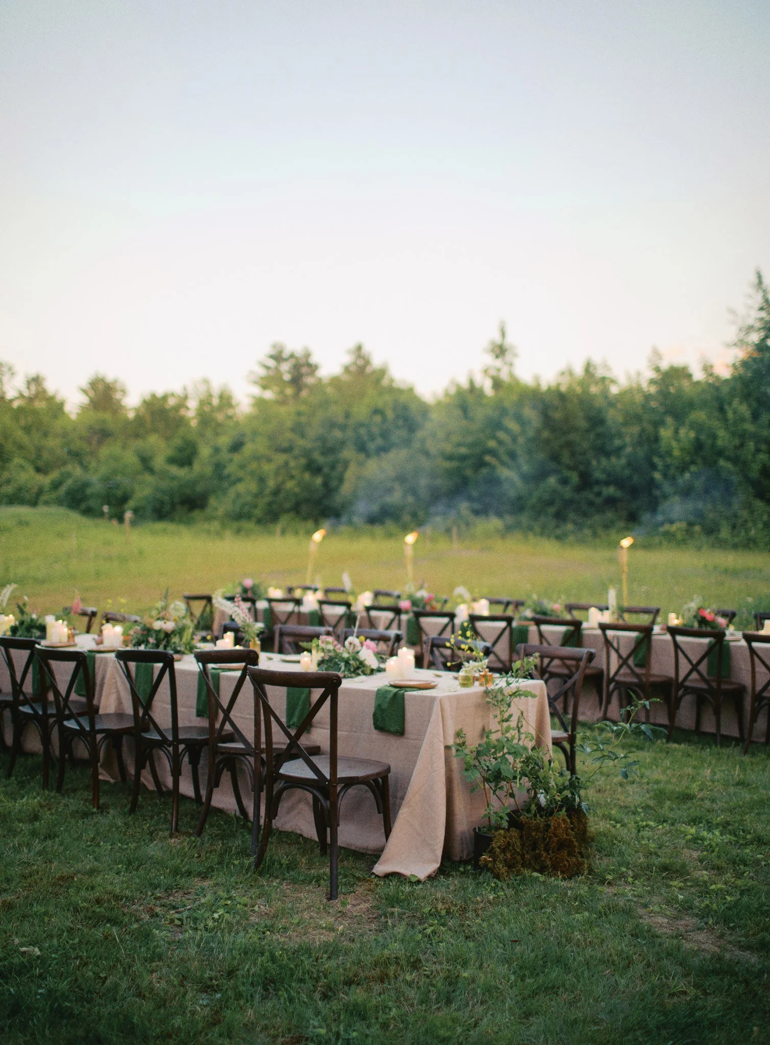 Outdoor event setup with tables, chairs, and candles on a grassy area, set against a backdrop of trees during dusk.