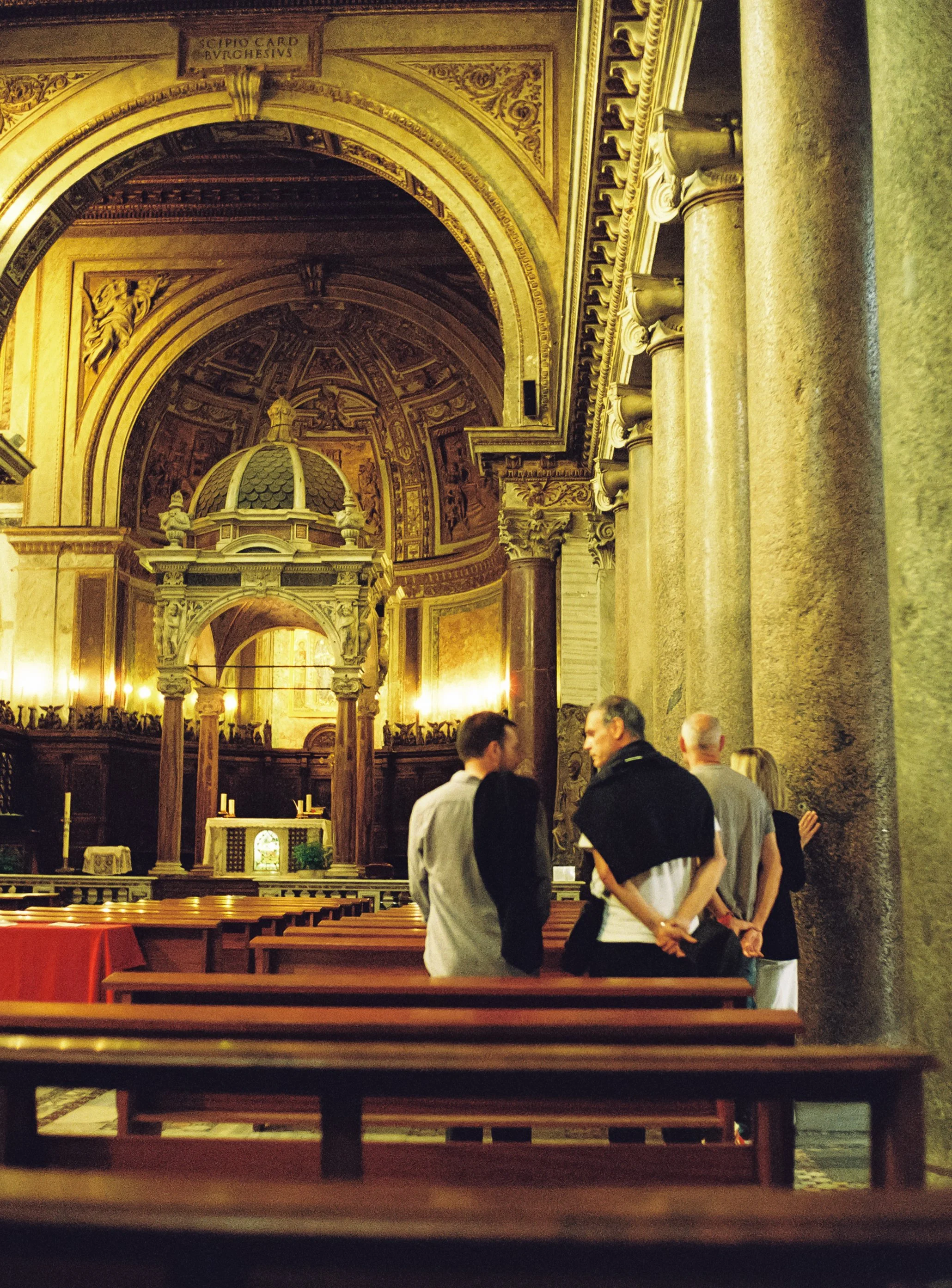 Four people are standing and talking inside a grand, ornate cathedral with gold accents, large columns, and elaborate ceiling artwork. Wooden pews and an altar are visible in the background.