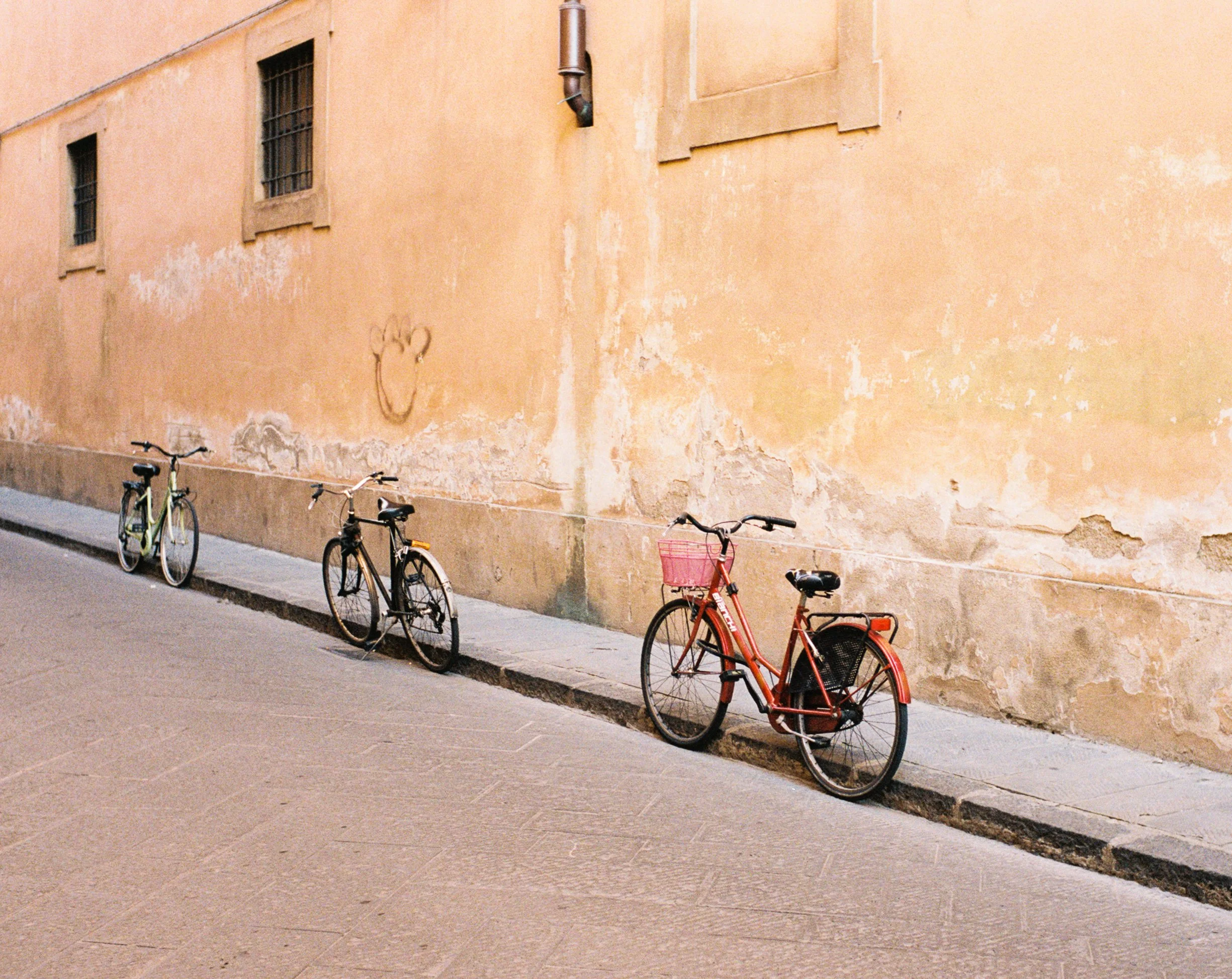 Three bicycles parked along a sidewalk next to an aged orange wall with small windows and graffiti.