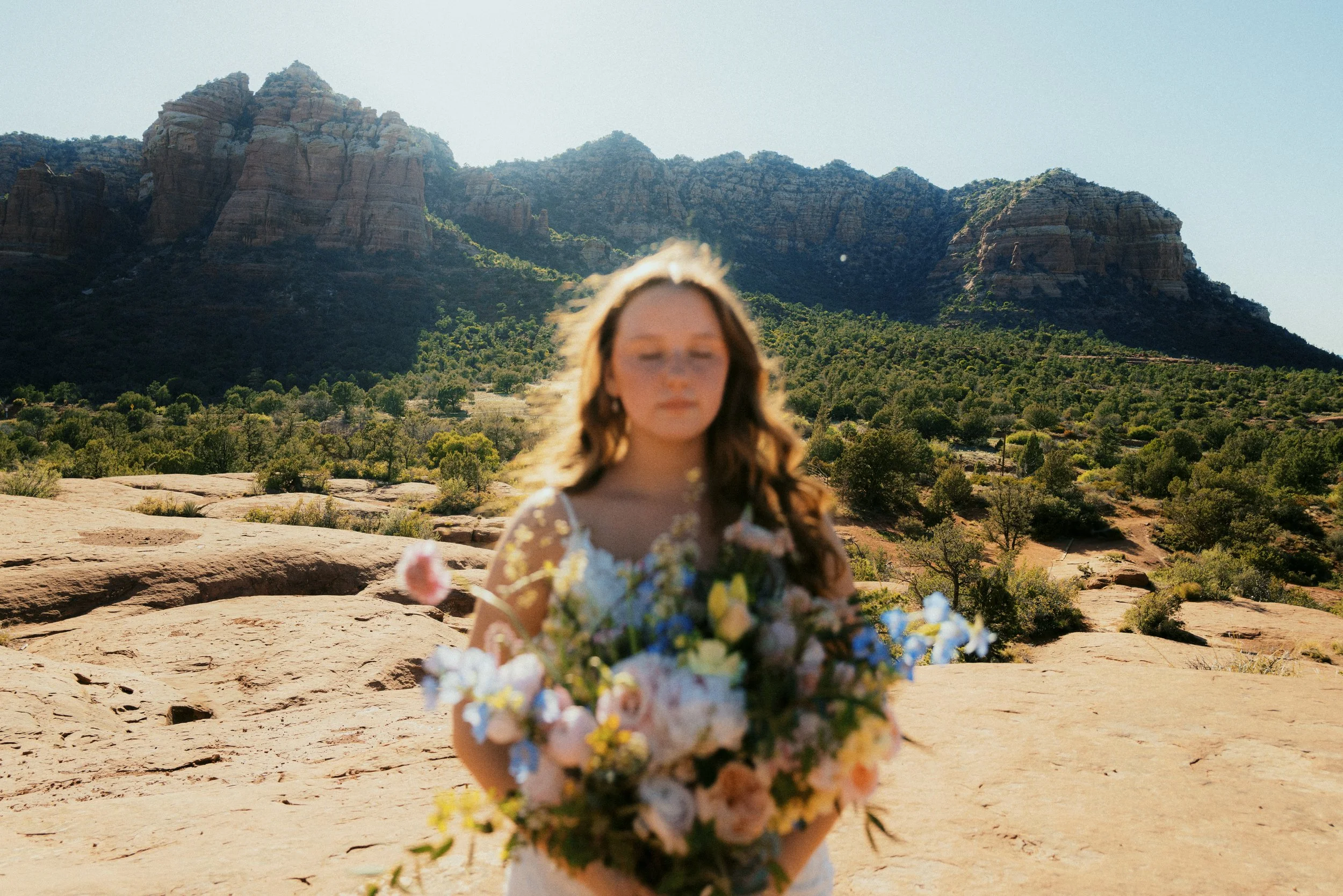 A young woman in a floral dress holding a bouquet of flowers outdoors with desert mountains and sparse greenery in the background.