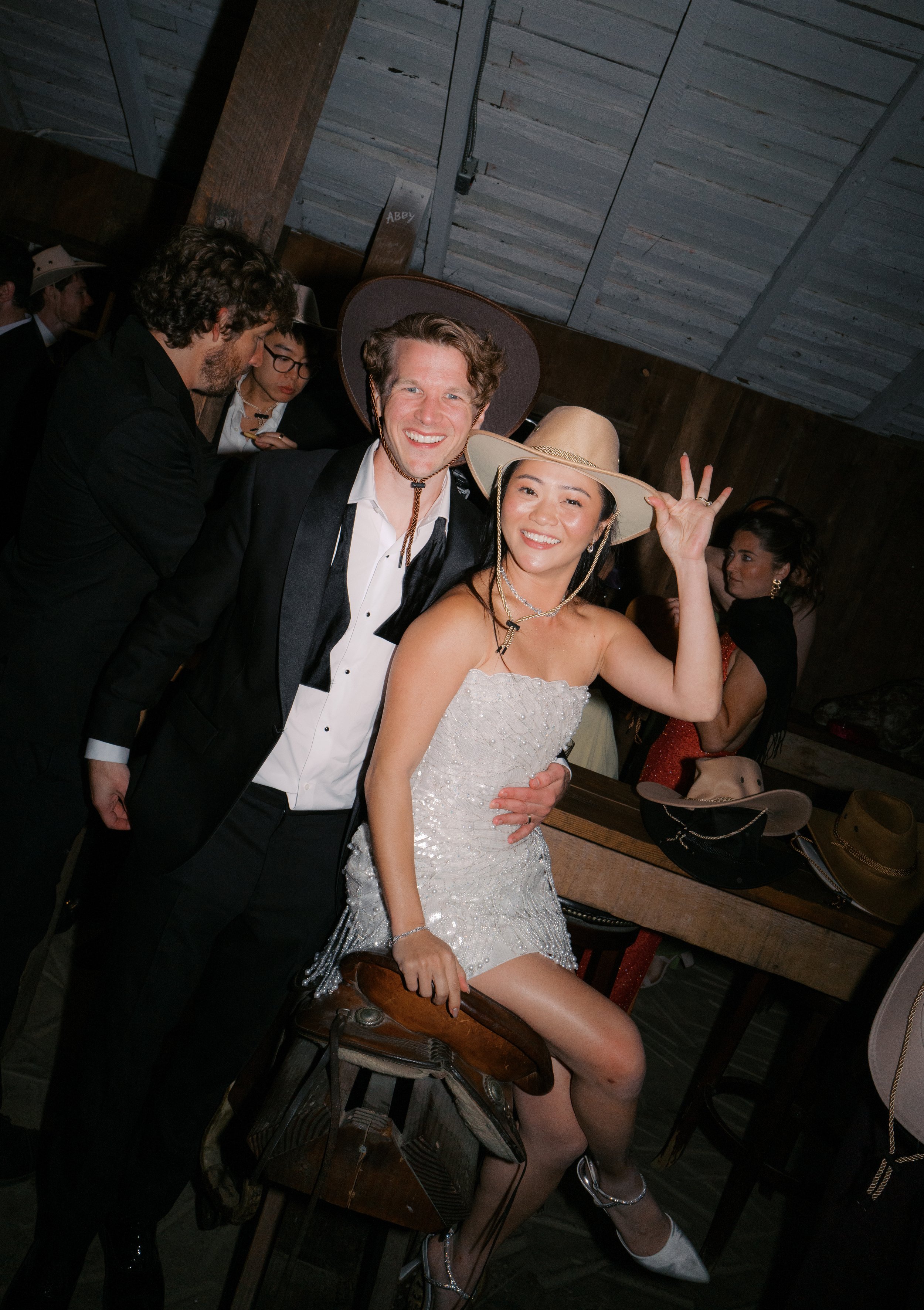 A group of people dressed in formal and costume attire at a party or social gathering, with one woman in a white dress and cowboy hat sitting on a wooden bar stool, making a peace sign, surrounded by men in suits and hats, in a rustic indoor setting.