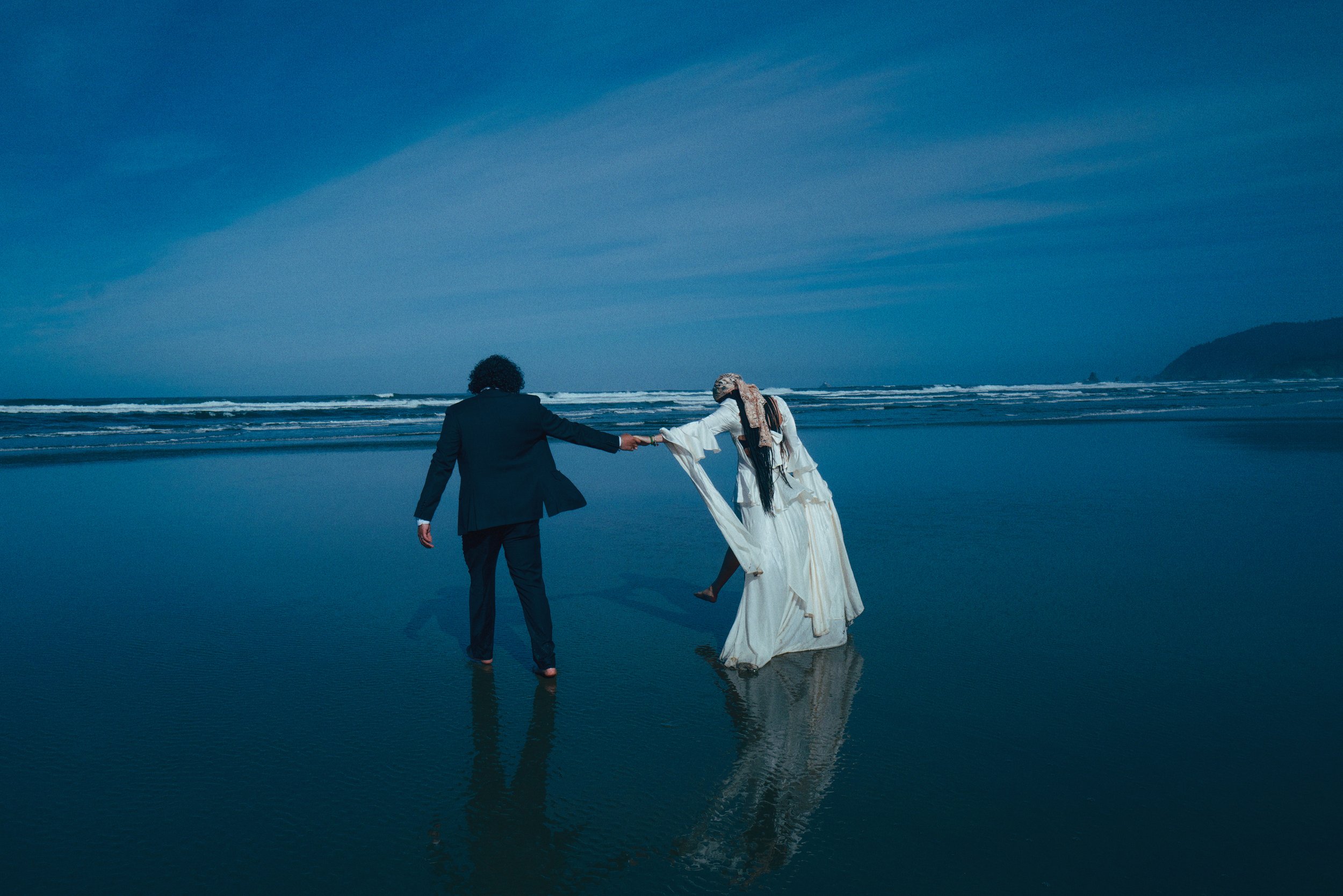 a bride and groom holding hands as they kick through the sand on cannon beach, oregon