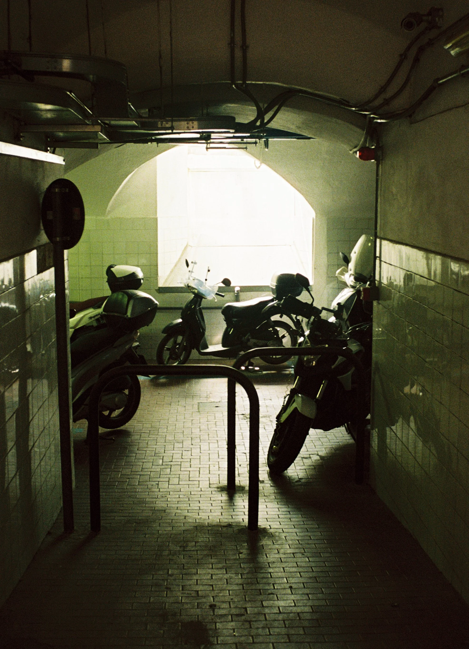 Underground parking area with four motorcycles parked near a metal railing, illuminated by natural light from an opening in the background.