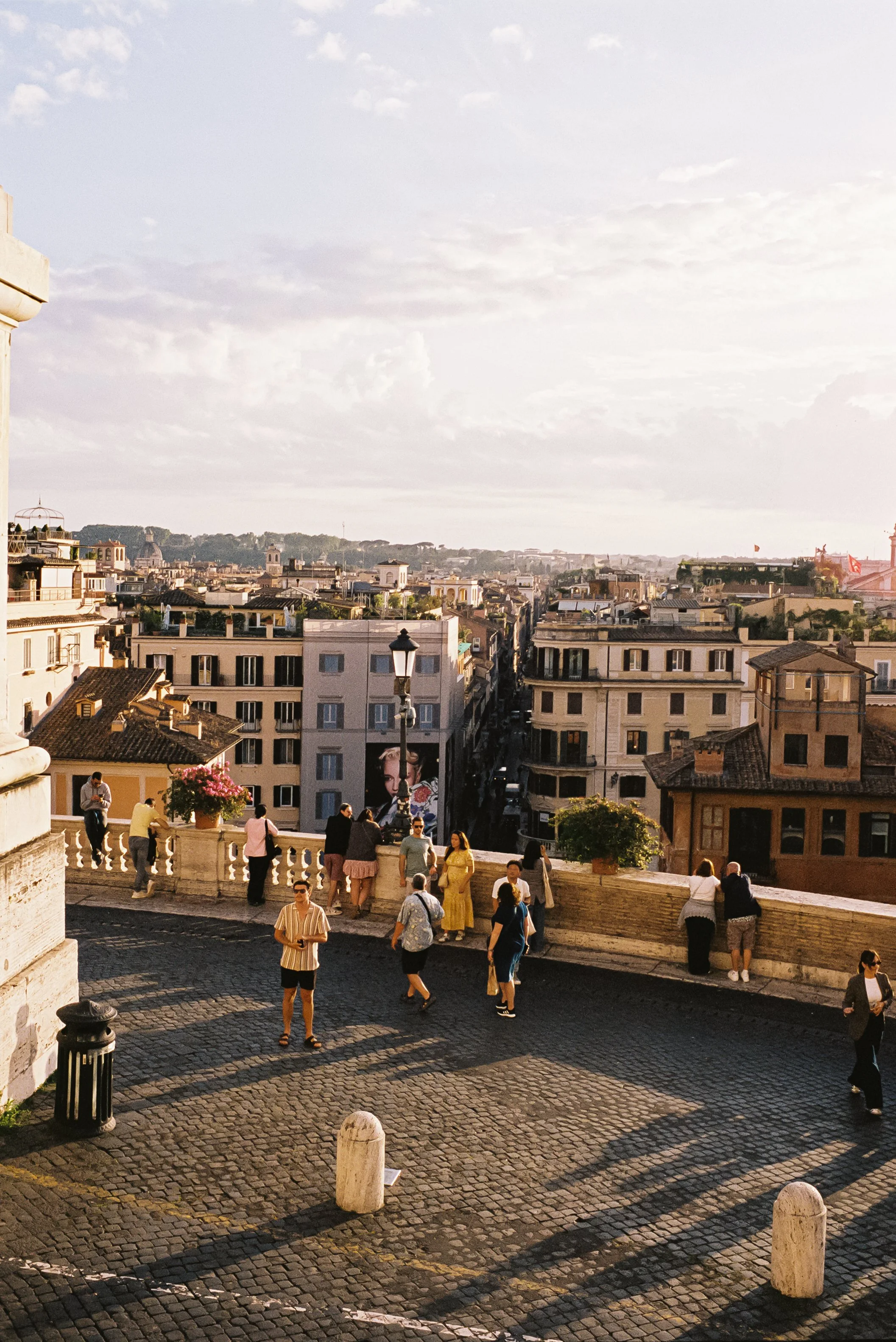 People gathered on a cobblestone street with a cityscape of buildings in the background, under a partly cloudy sky during daytime.