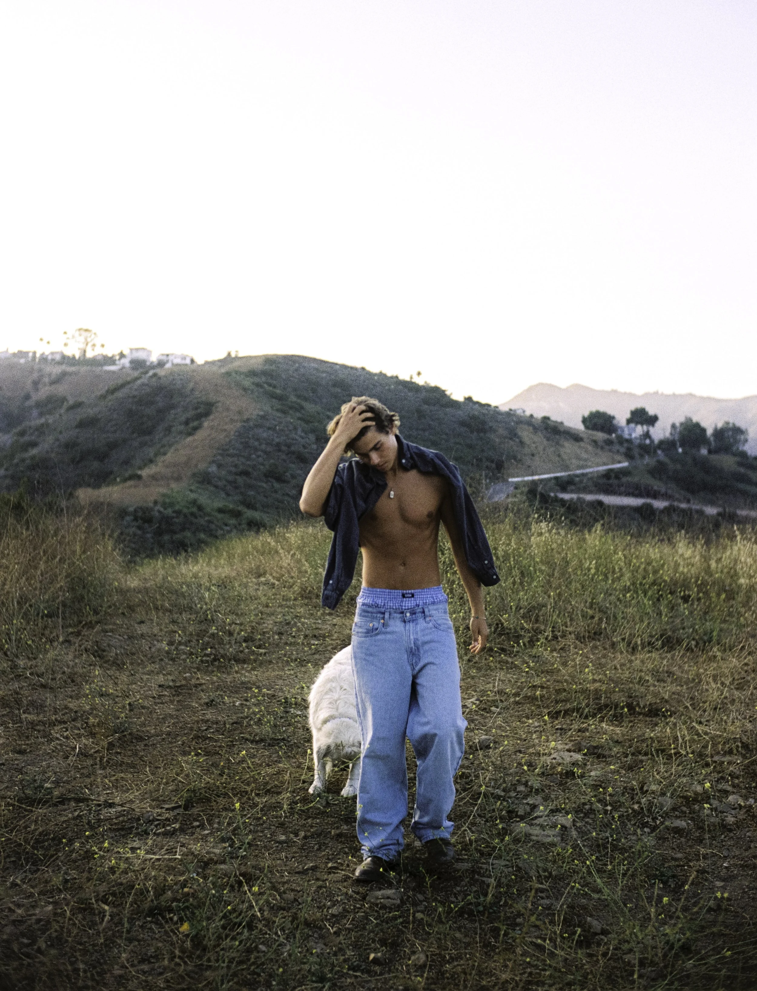 A young man walking through a grassy field with hills in the background during sunset, shirtless with an open jacket and wearing baggy jeans.