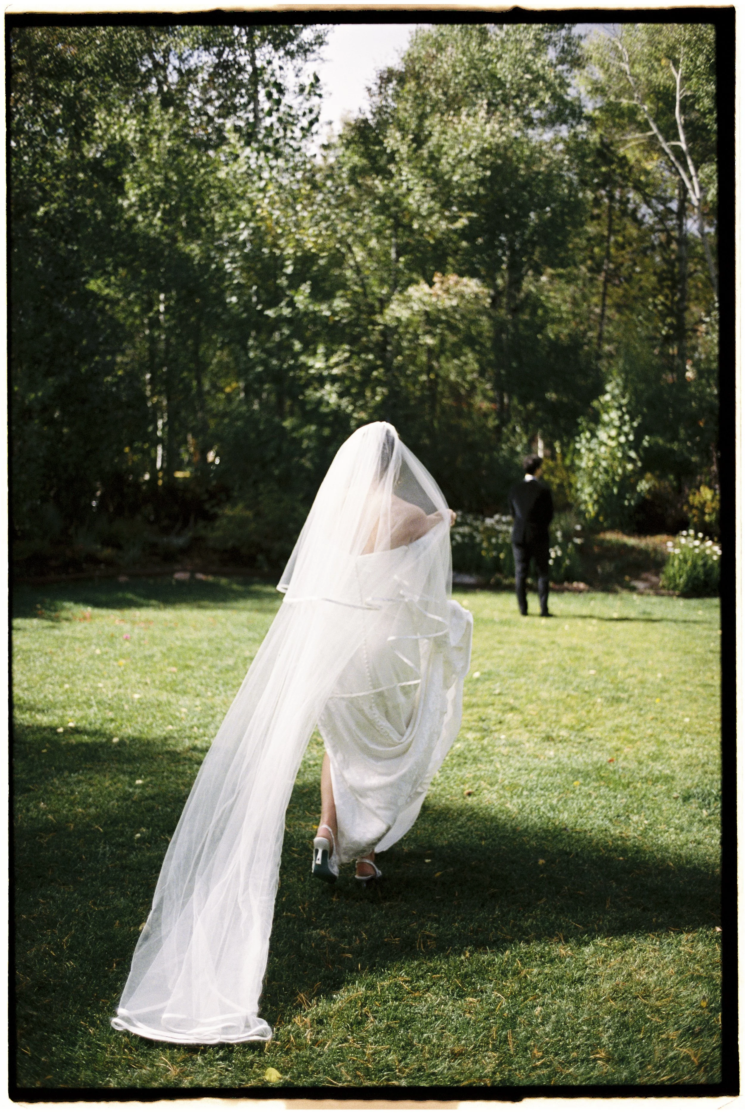 A bride in a wedding dress walking in a garden with a veil flowing behind her, and a man in a suit standing in the background under trees.