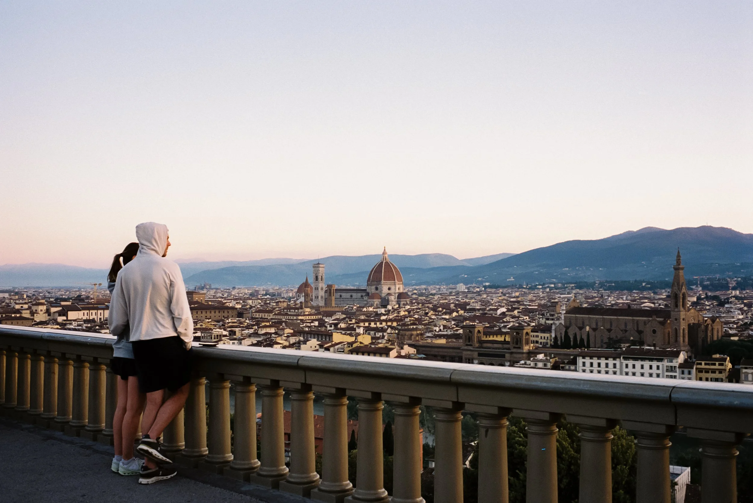 Two people watch over Florence from a viewing platform, with the city skyline, the Florence Cathedral's dome, and the mountains in the background.