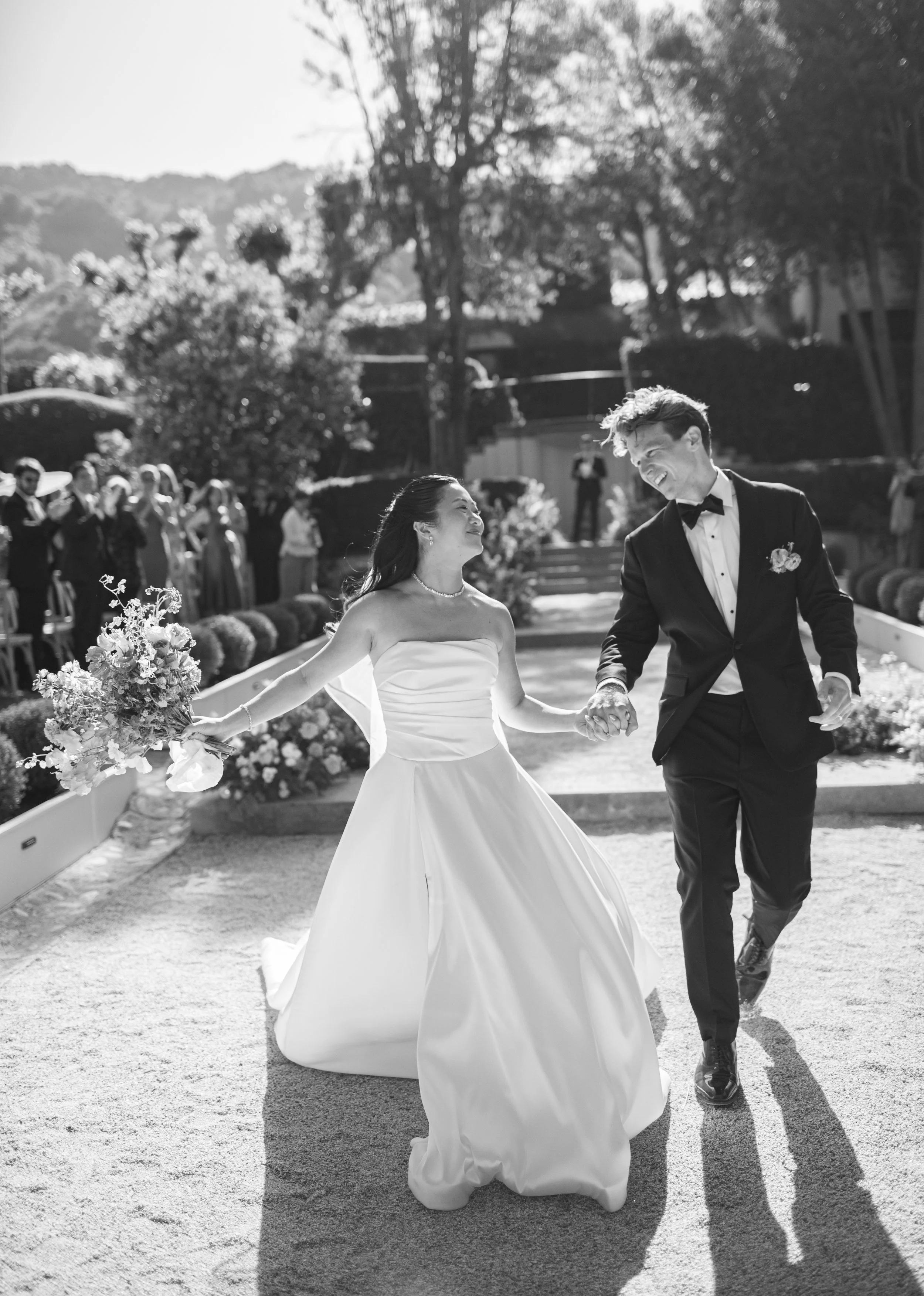 A black and white photo of a bride and groom holding hands and dancing at their wedding outdoors, with guests in the background watching and trees and a house in the distance.