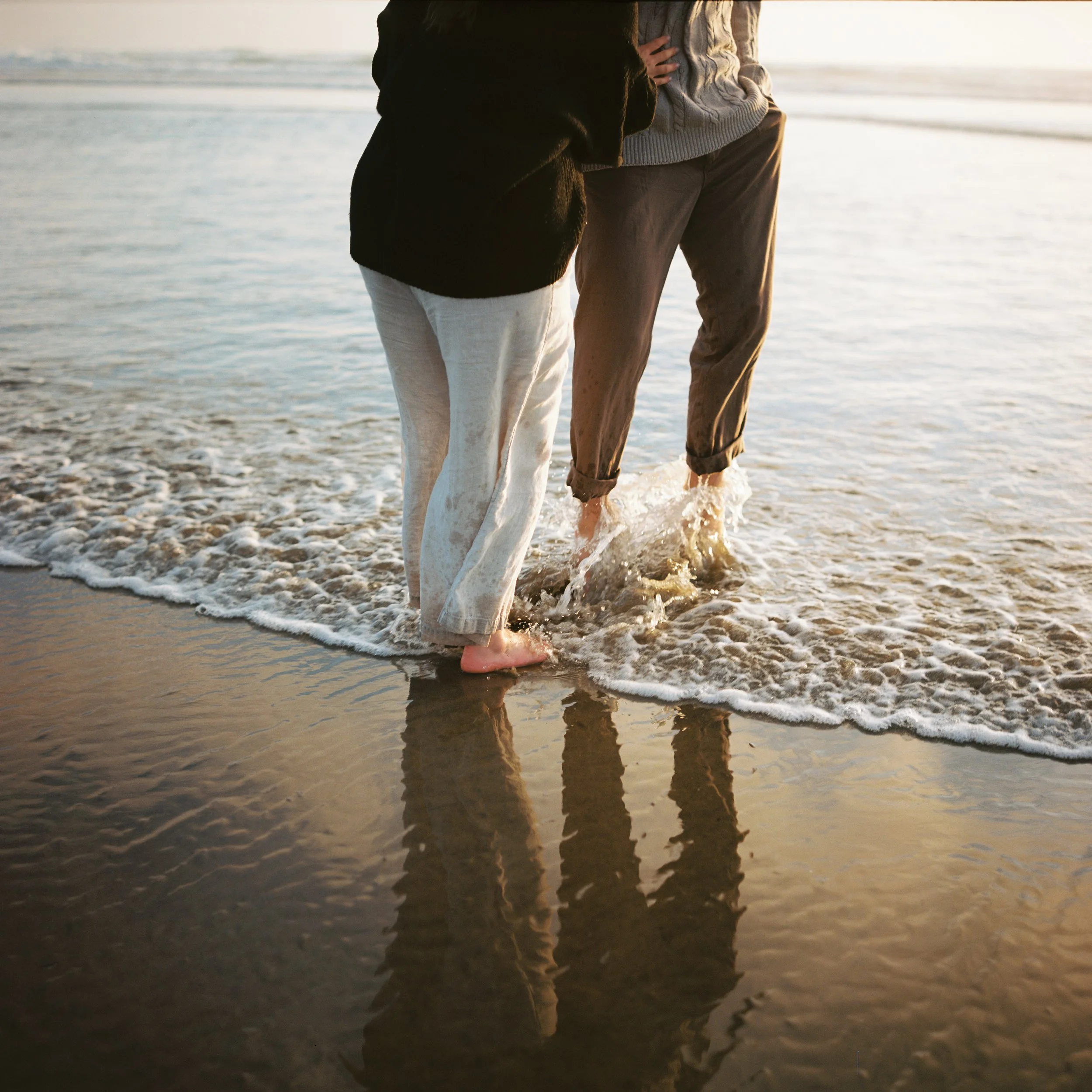 bride and groom dipping toes into the ocean as a wave crashes on their feet