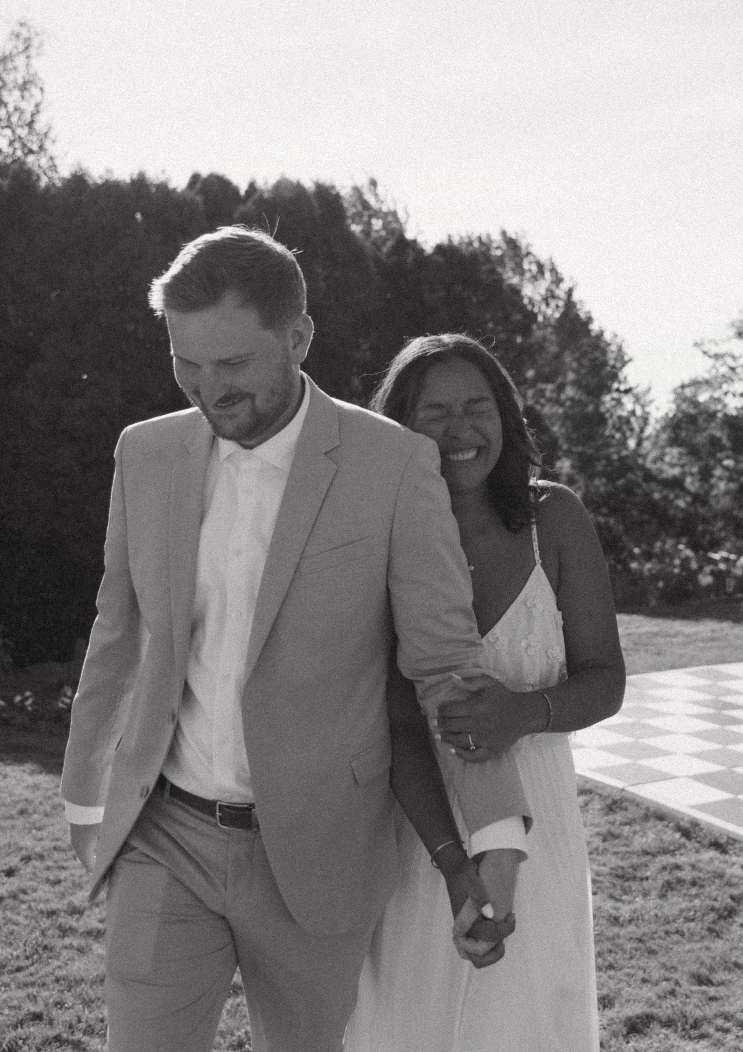 A black-and-white photo of a smiling woman and a man holding hands outdoors, with trees and a checkered floor in the background.