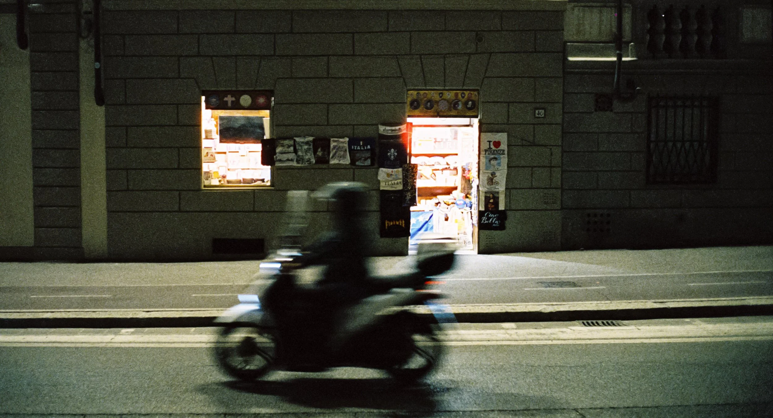 A person riding a scooter on a city street at night in front of a lit storefront with signs and posters on the door and window.