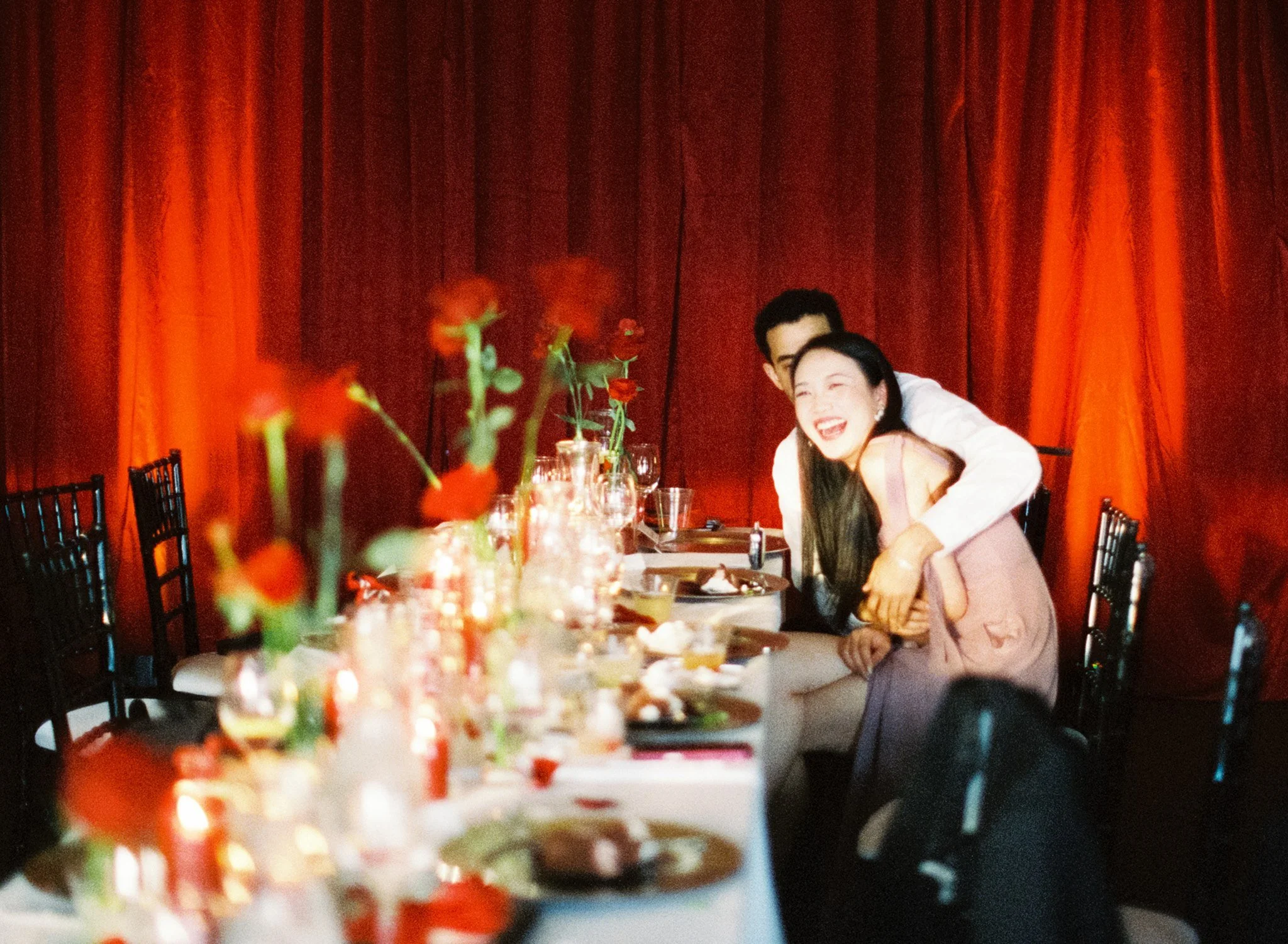 a couple laughing at the camera on a table set with roses and red tablescapes with red curtains