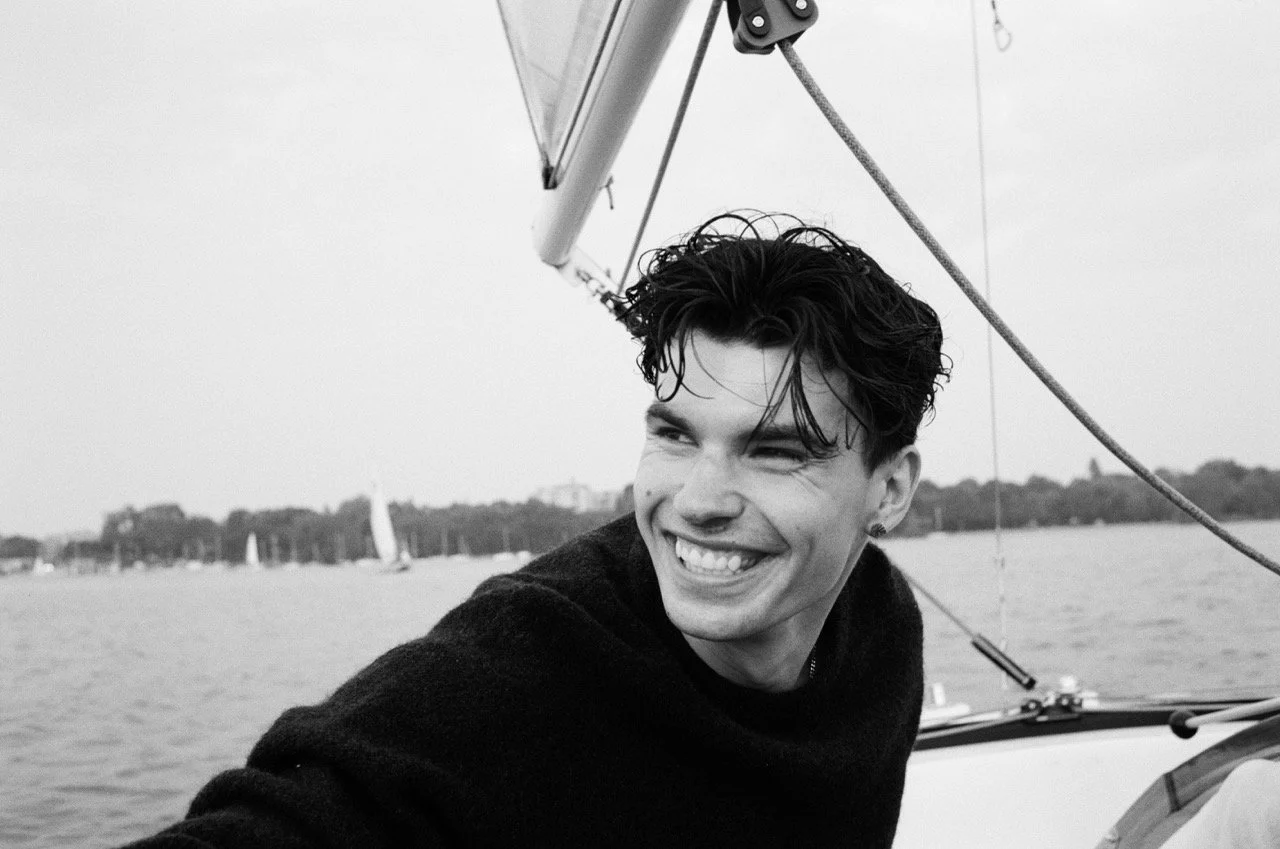 A young man with dark, wavy hair smiling on a boat, with water and sailboats in the background, in black and white.