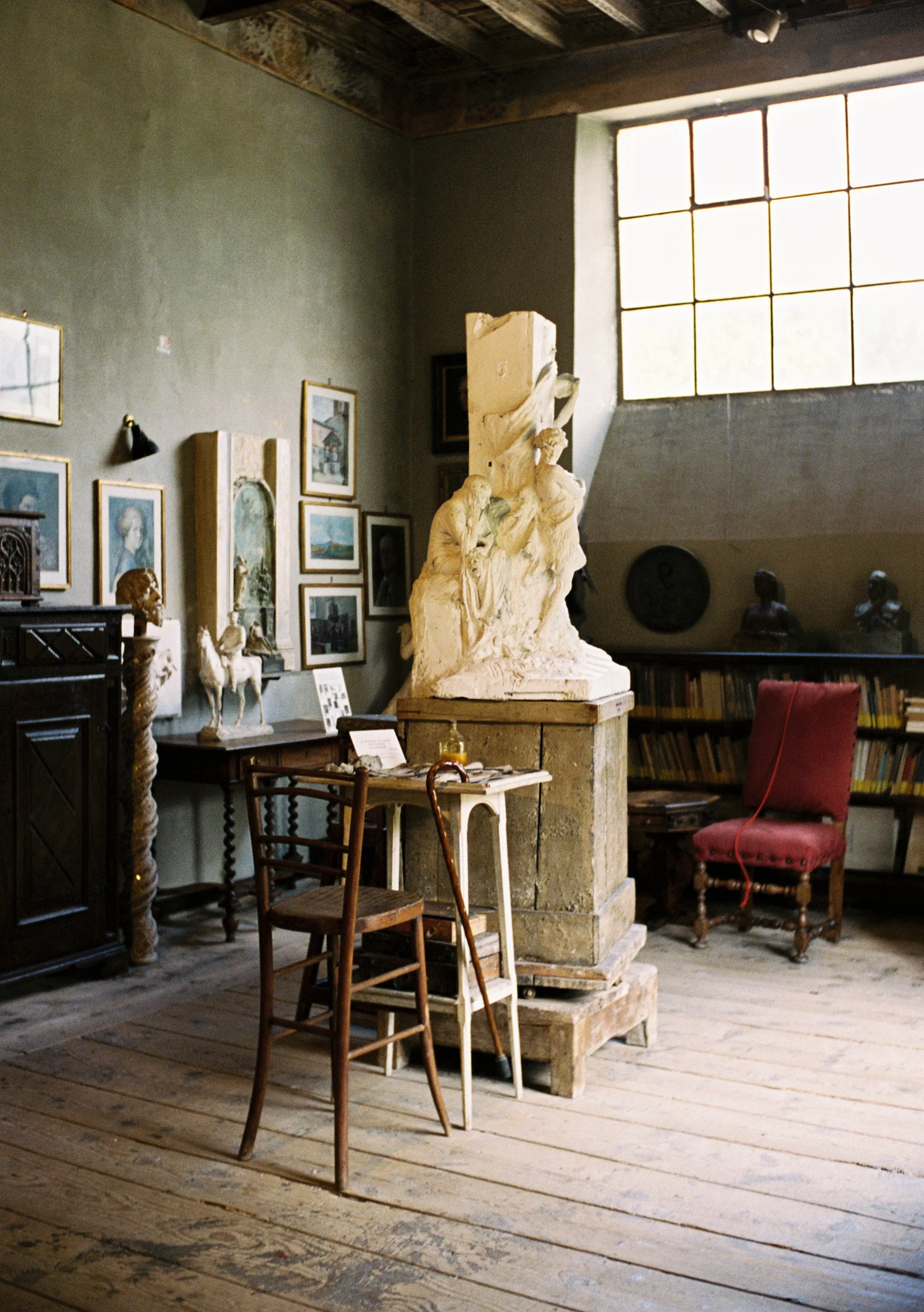 Room with art and sculpture, including a large white sculpture on a pedestal, framed pictures on the wall, wooden chairs, and shelves with books and statues, lit by a large window.