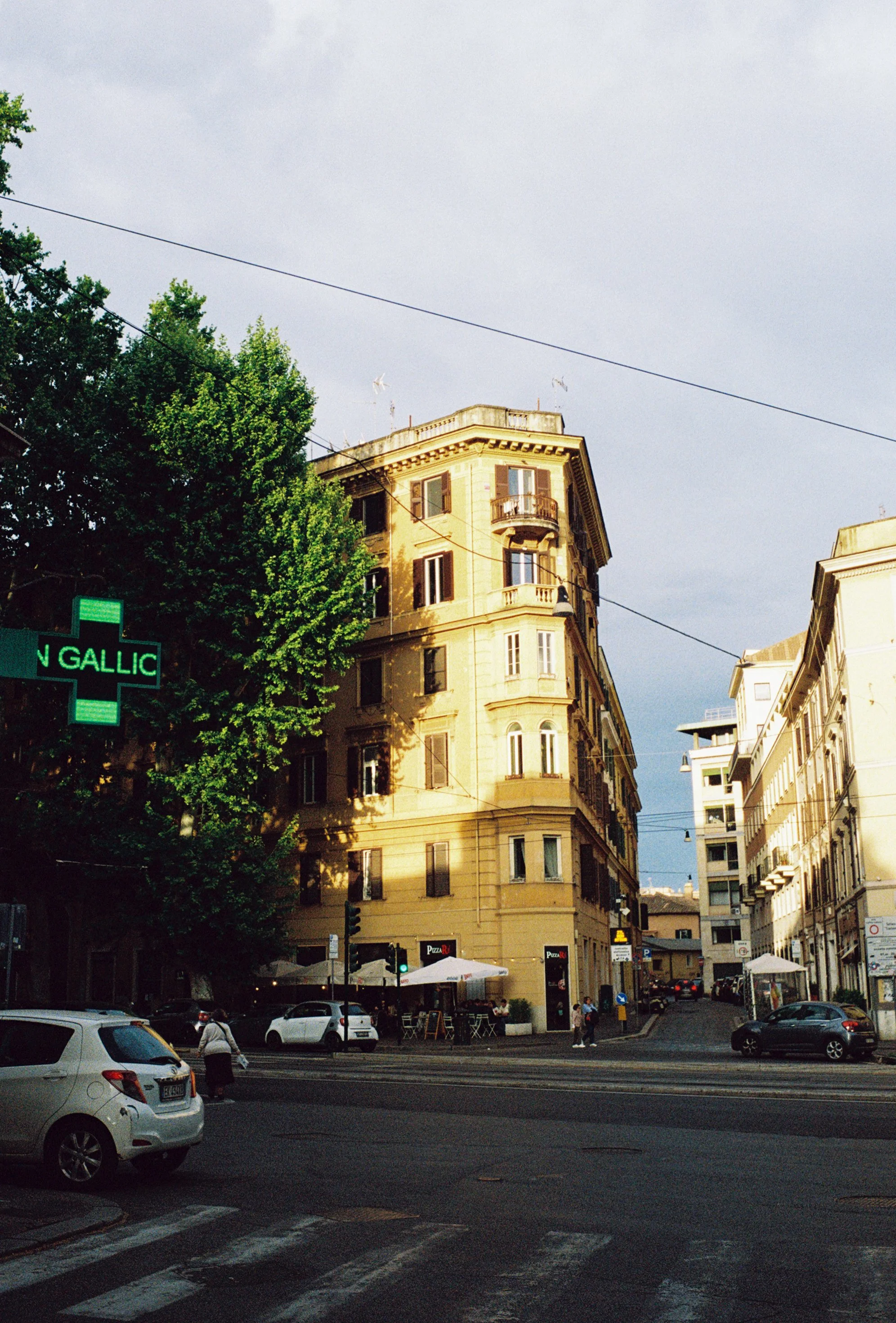 Street scene in an urban area with a large yellow building, cars on the road, and pedestrians walking. There is a green cross sign on a pole, and outdoor seating for a restaurant or cafe.