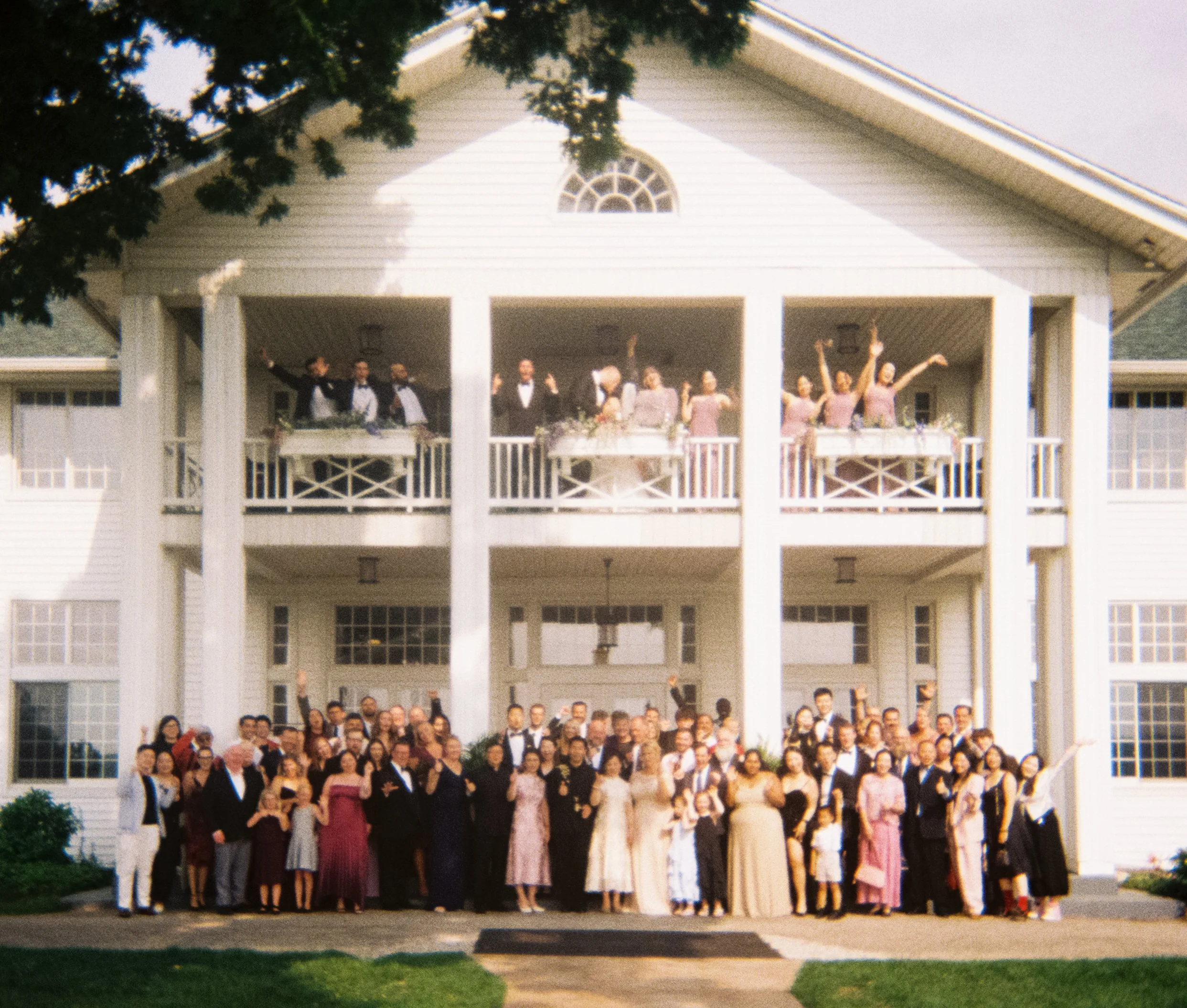 A large group of people in formal attire standing on the front lawn of a white house, possibly at a wedding or formal celebration, with trees and a pathway visible.