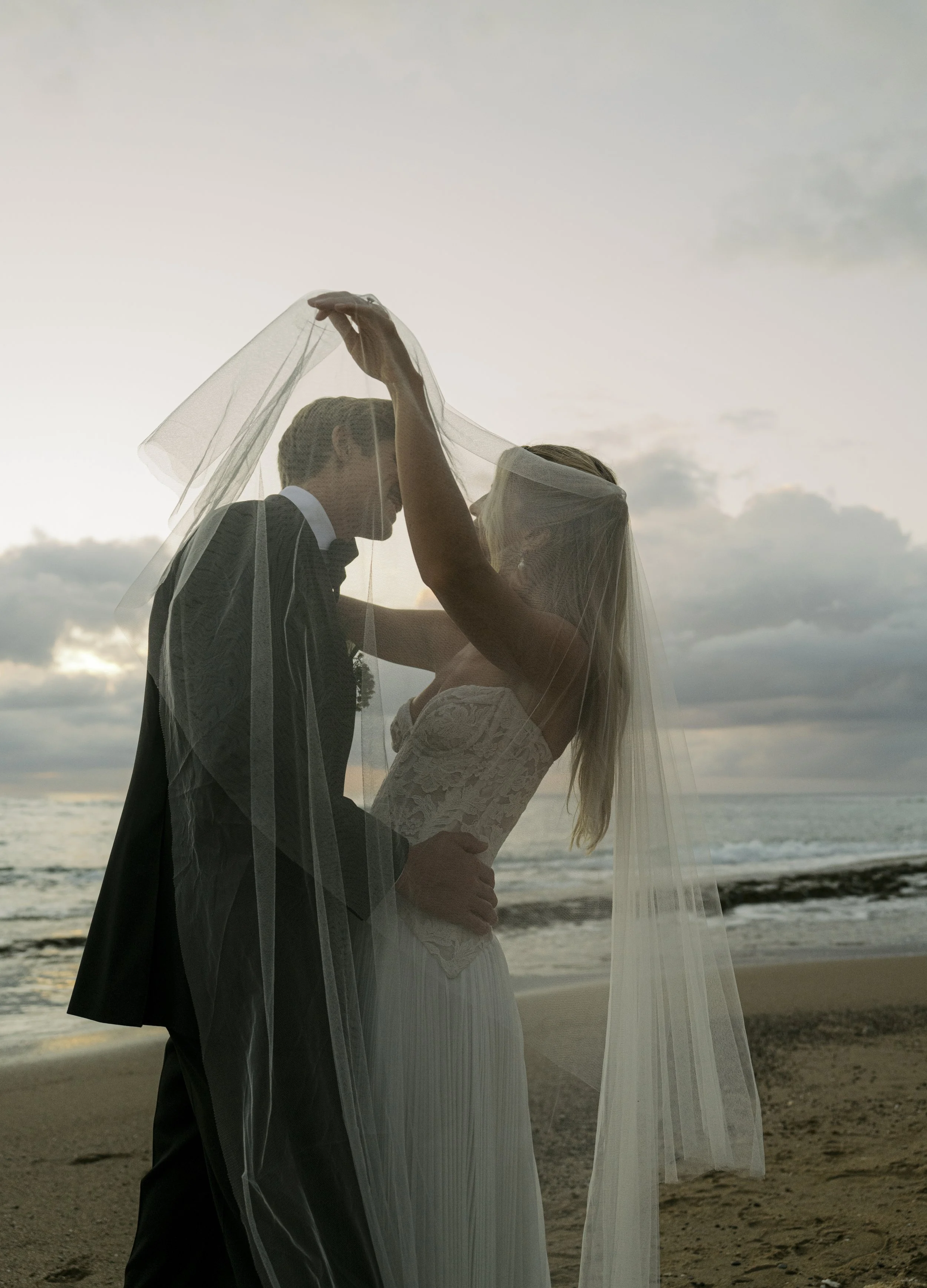 A bride and groom stand close together on the beach at sunset, sharing an intimate moment under a veil held by the bride's arm.