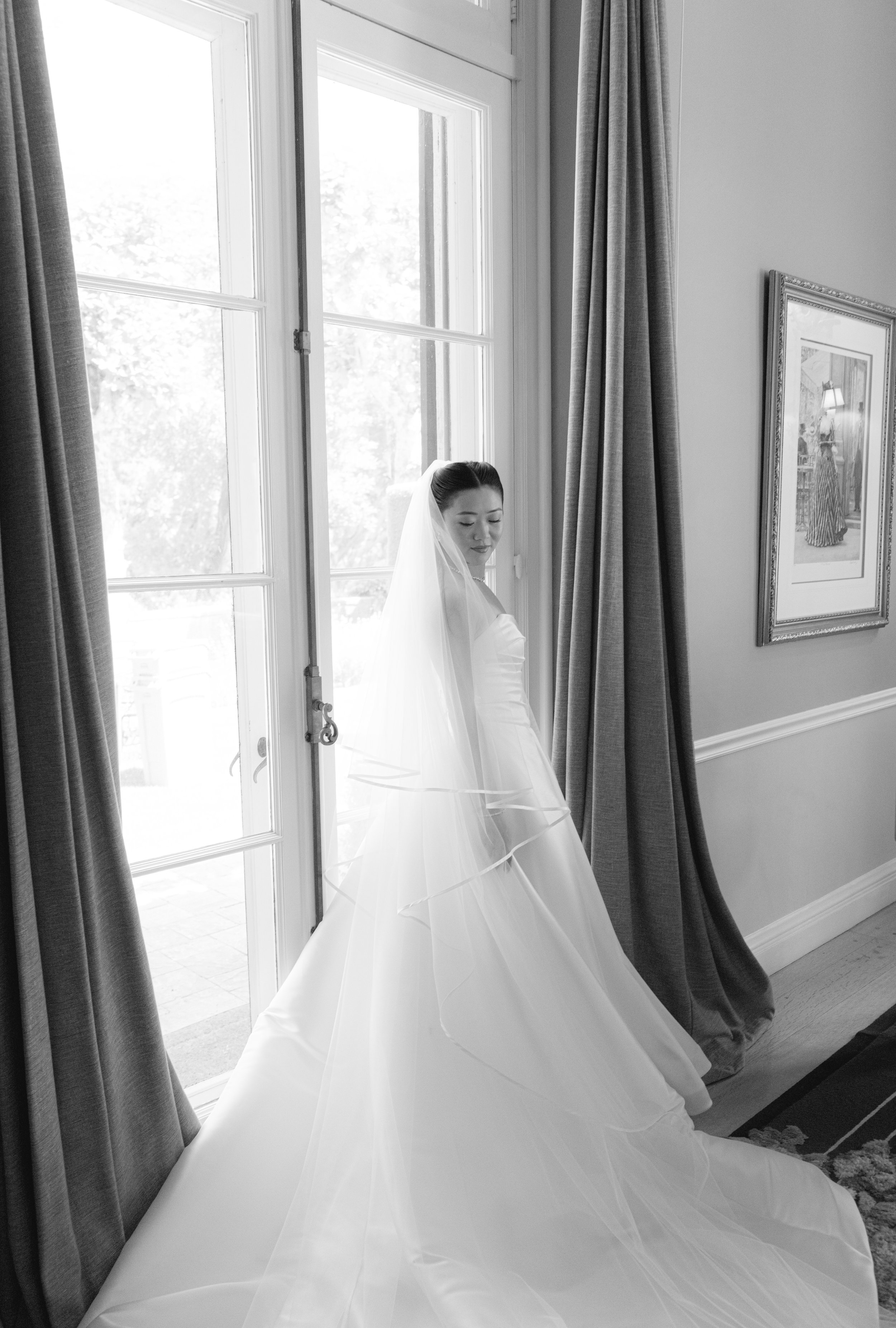 A bride in a wedding dress with a veil, standing inside near a large window with curtains, looking down, with a framed picture on the wall behind her.