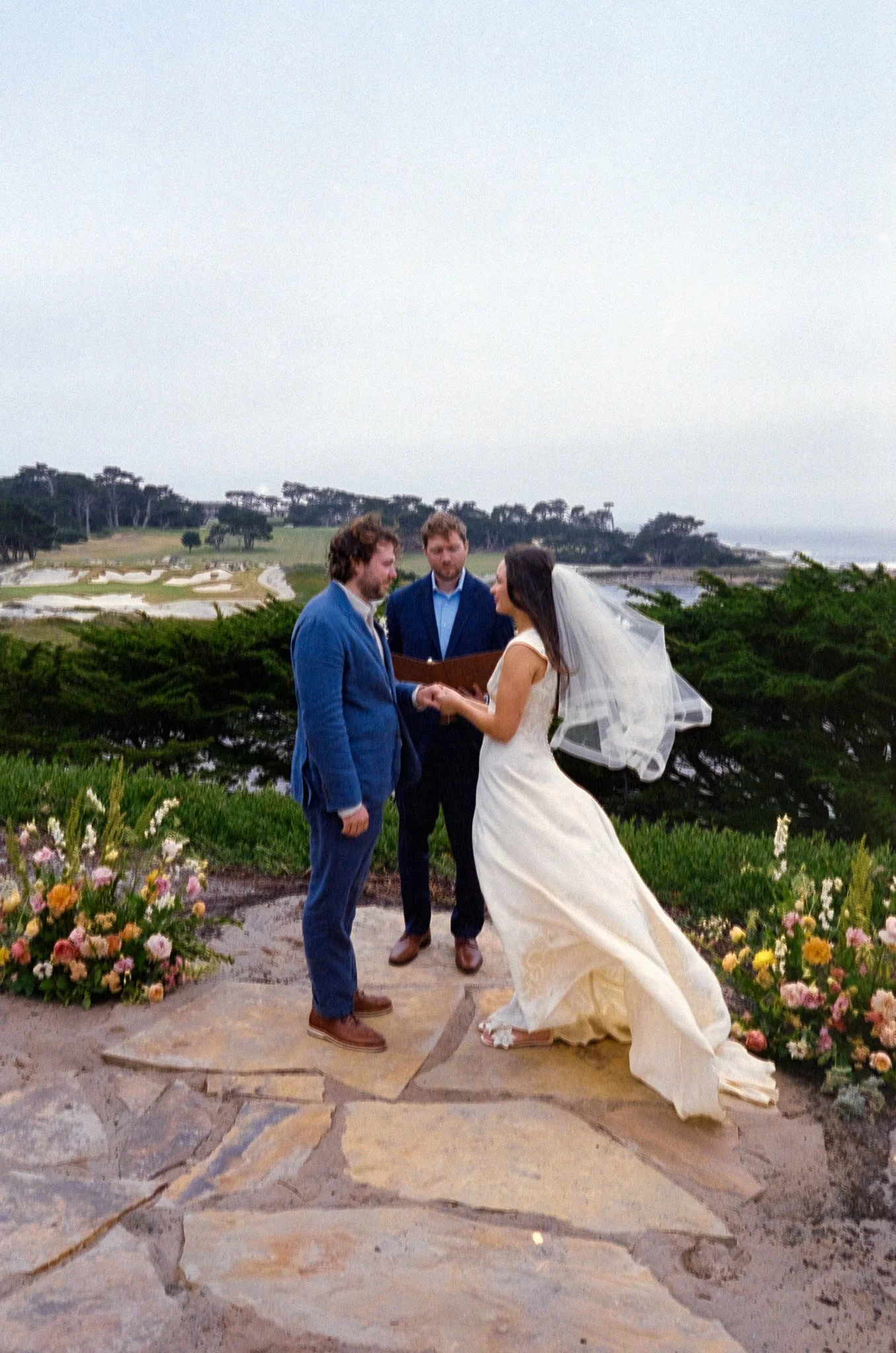 A wedding ceremony outdoors with a bride and groom holding hands, standing on a stone path near flower arrangements and greenery, with an officiant and a coastal landscape in the background.