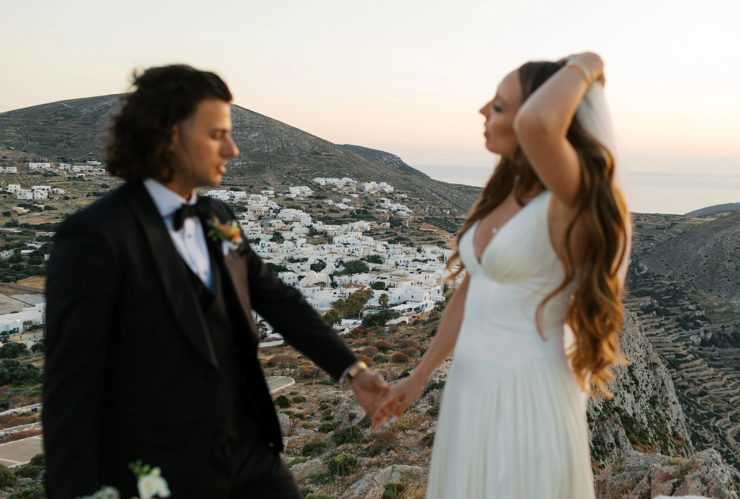 A couple dressed in wedding attire holding hands on a hillside with white buildings and hills in the background during sunset.