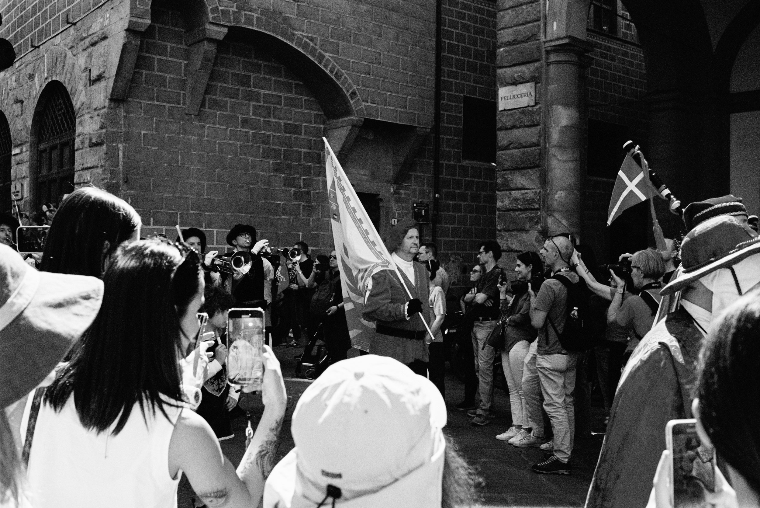Crowd gathered on a street holding flags and cameras, with a brick building in the background.