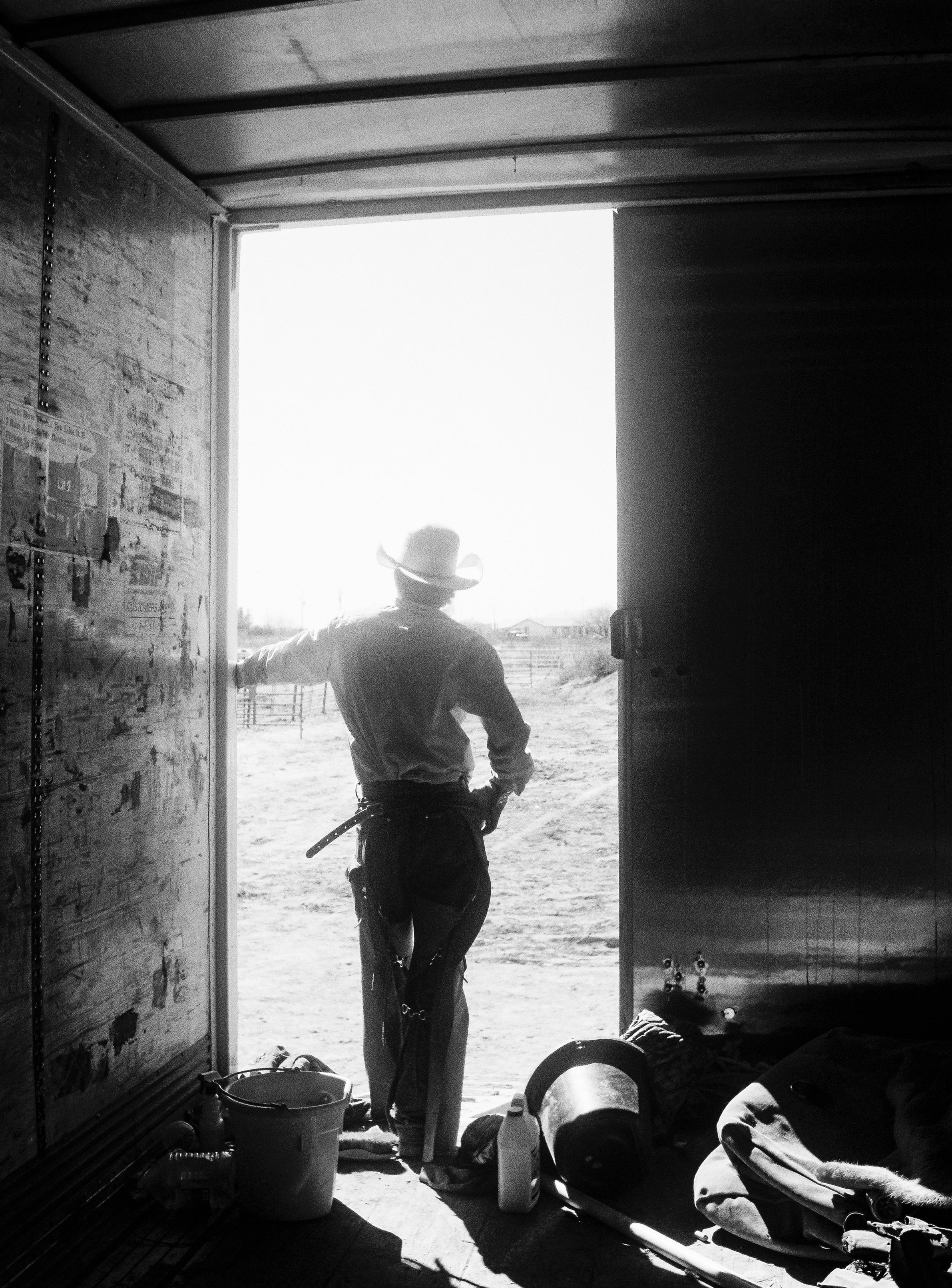A cowboy in a hat standing in a barn doorway, looking outside on a sunny day, with farming materials and equipment on the ground inside the barn.
