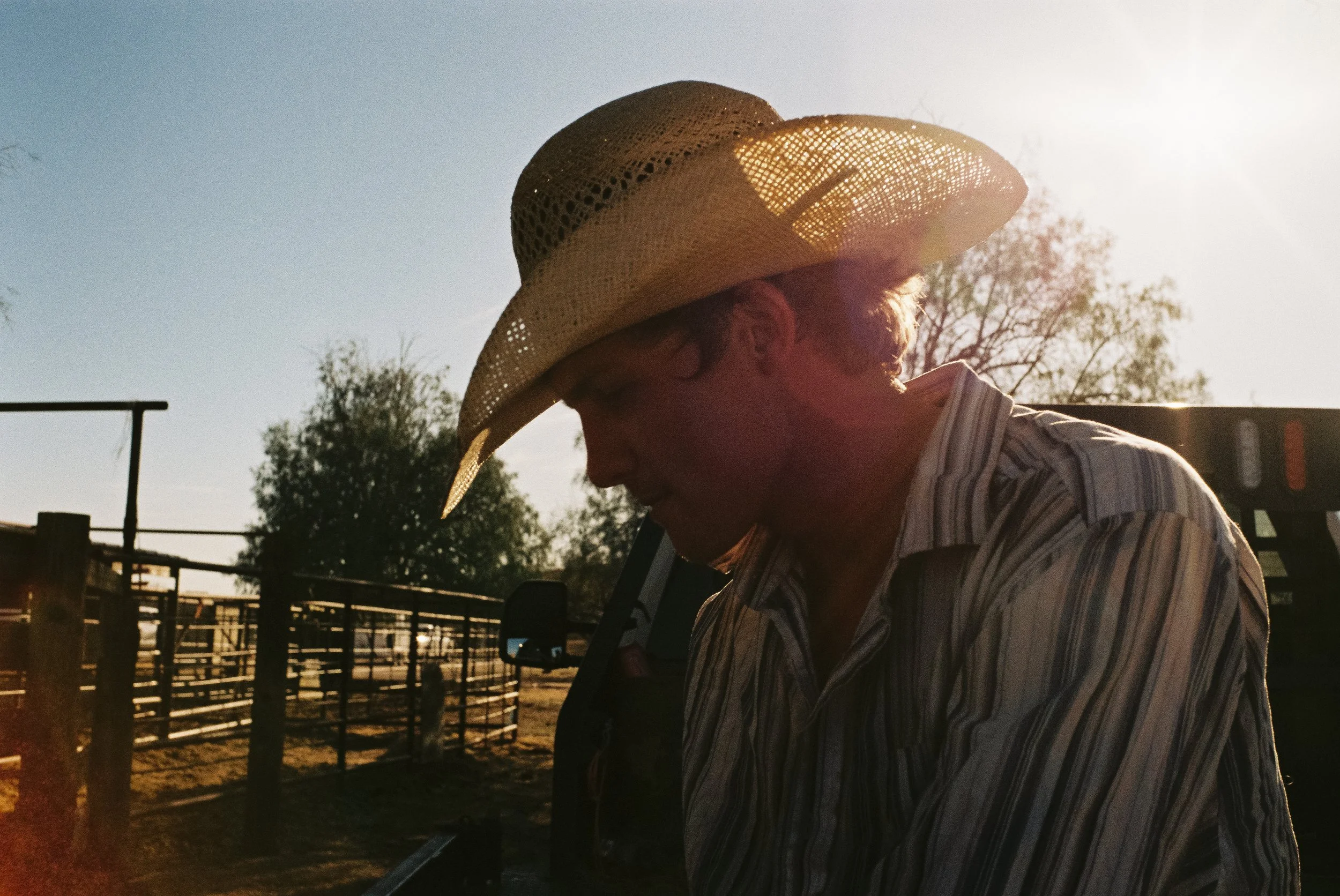A person wearing a cowboy hat and a striped shirt stands outdoors near a fence, with sunlight shining behind them.