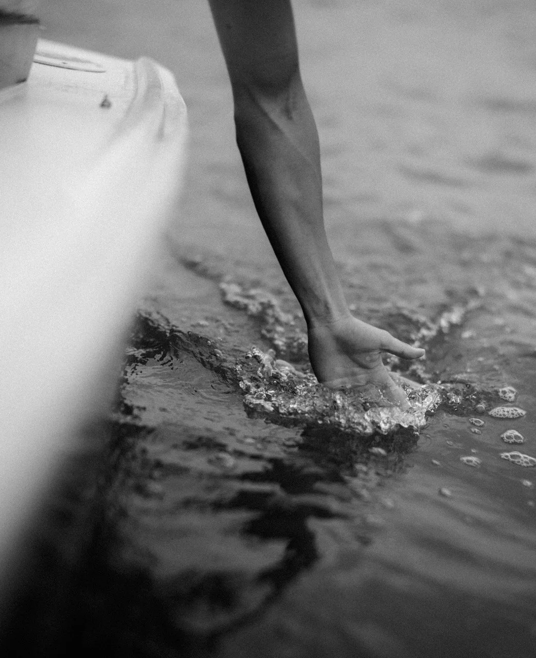 A person reaching into the water at the shoreline, with their hand submerged and creating ripples and small bubbles in the water.