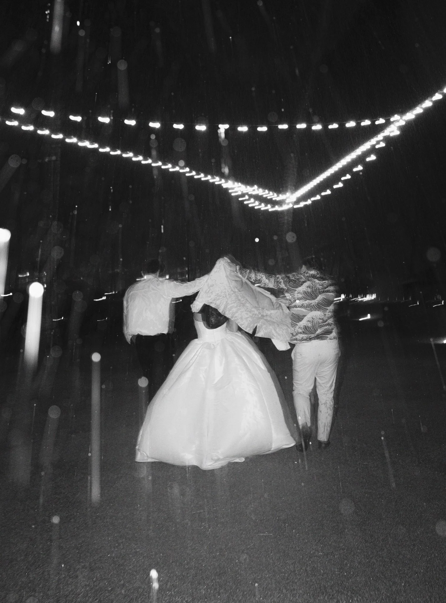 People dancing at night under string lights, wearing traditional clothing, with rain falling.