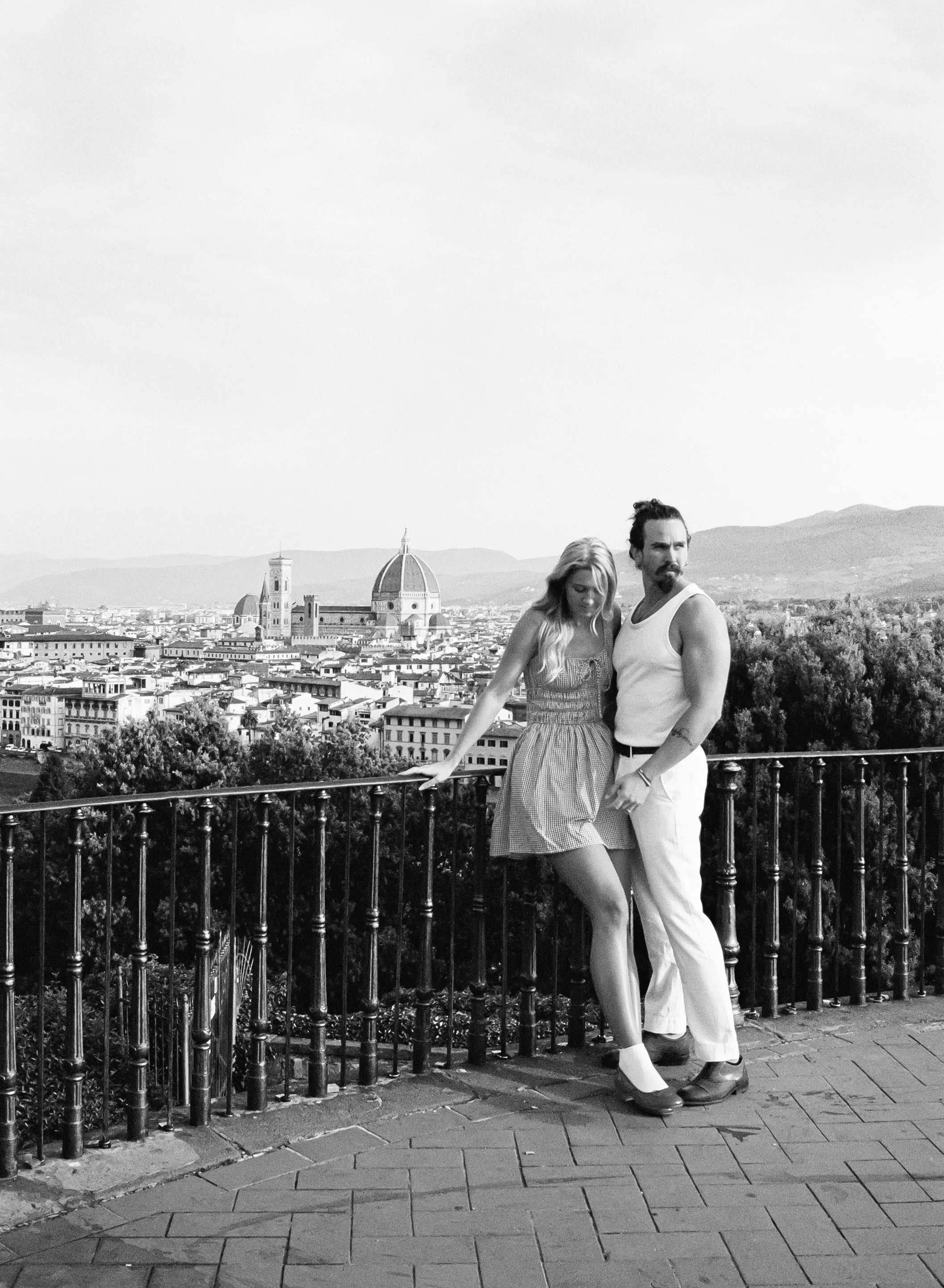 Black and white photo of a man and woman standing close to each other by a railing, with a cityscape of Florence, Italy, in the background, including the Florence Cathedral with its dome.