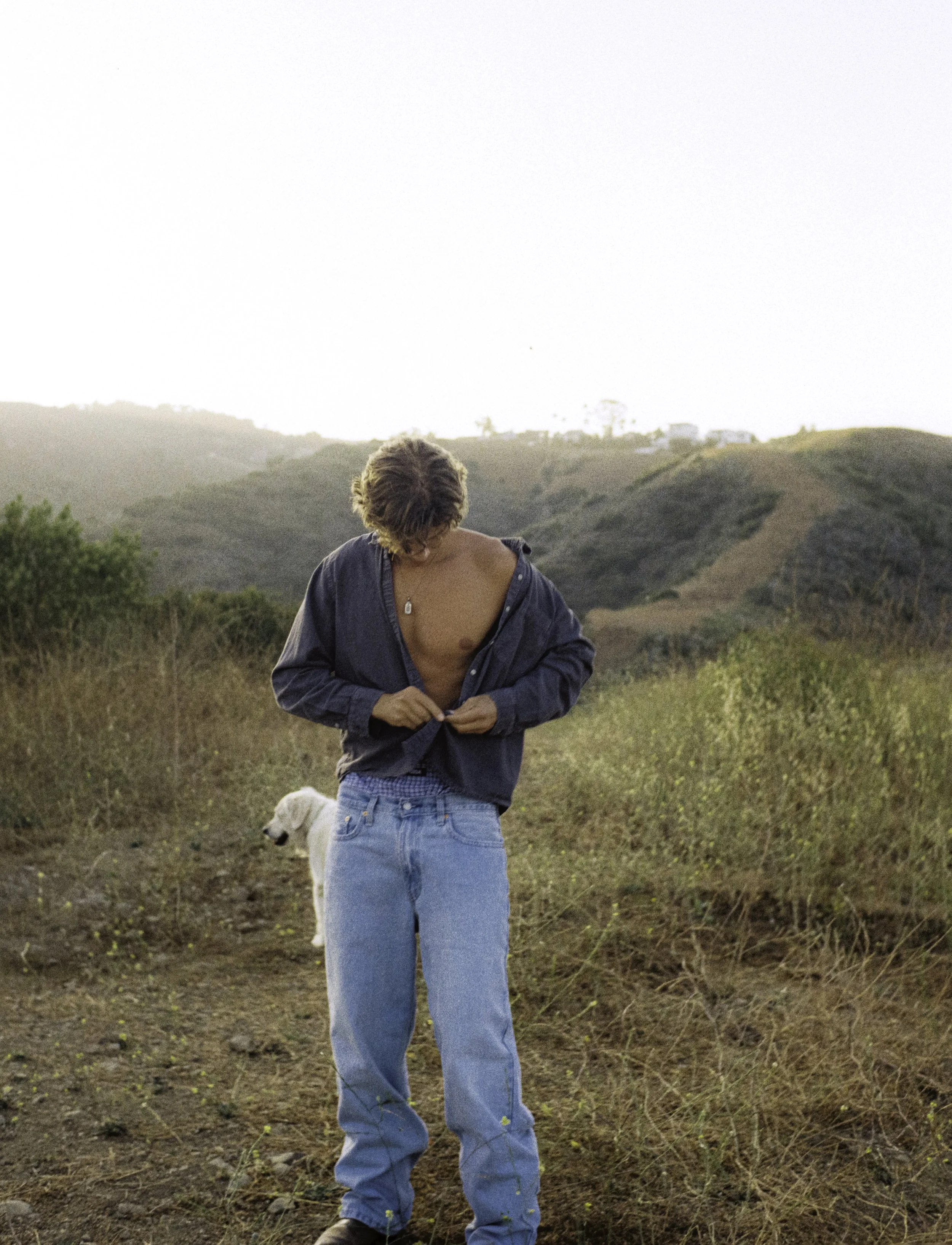 Person with curly hair partially unzipping a dark jacket, standing outdoors on a dirt trail with a dog in the background, with rolling hills and a bright sky in the distance.