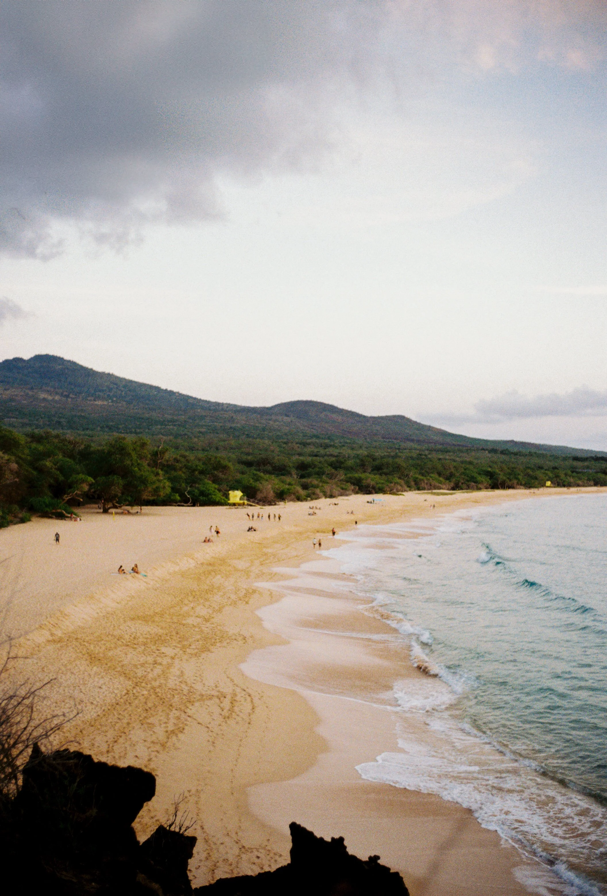 Beach with sandy shoreline, some people sitting and walking, green trees along the coast, hills in the background, cloudy sky.
